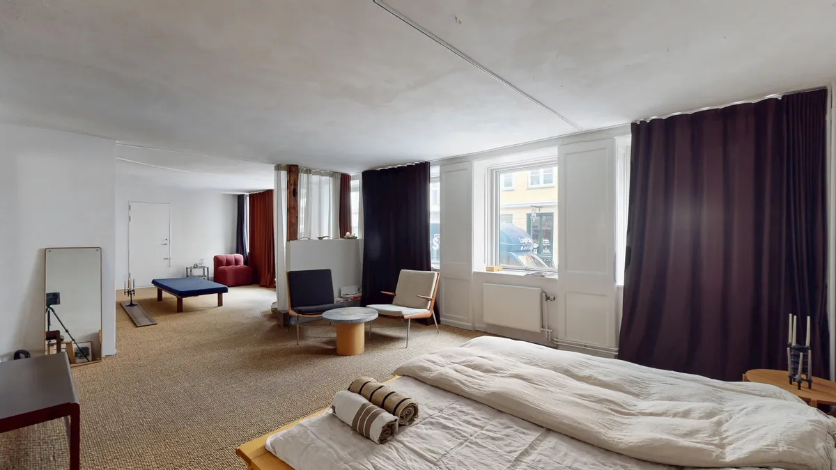 Elegant værelse med hvid væg og beige gulv samt moderne møbler, en høj seng med to puder og en stående spejl