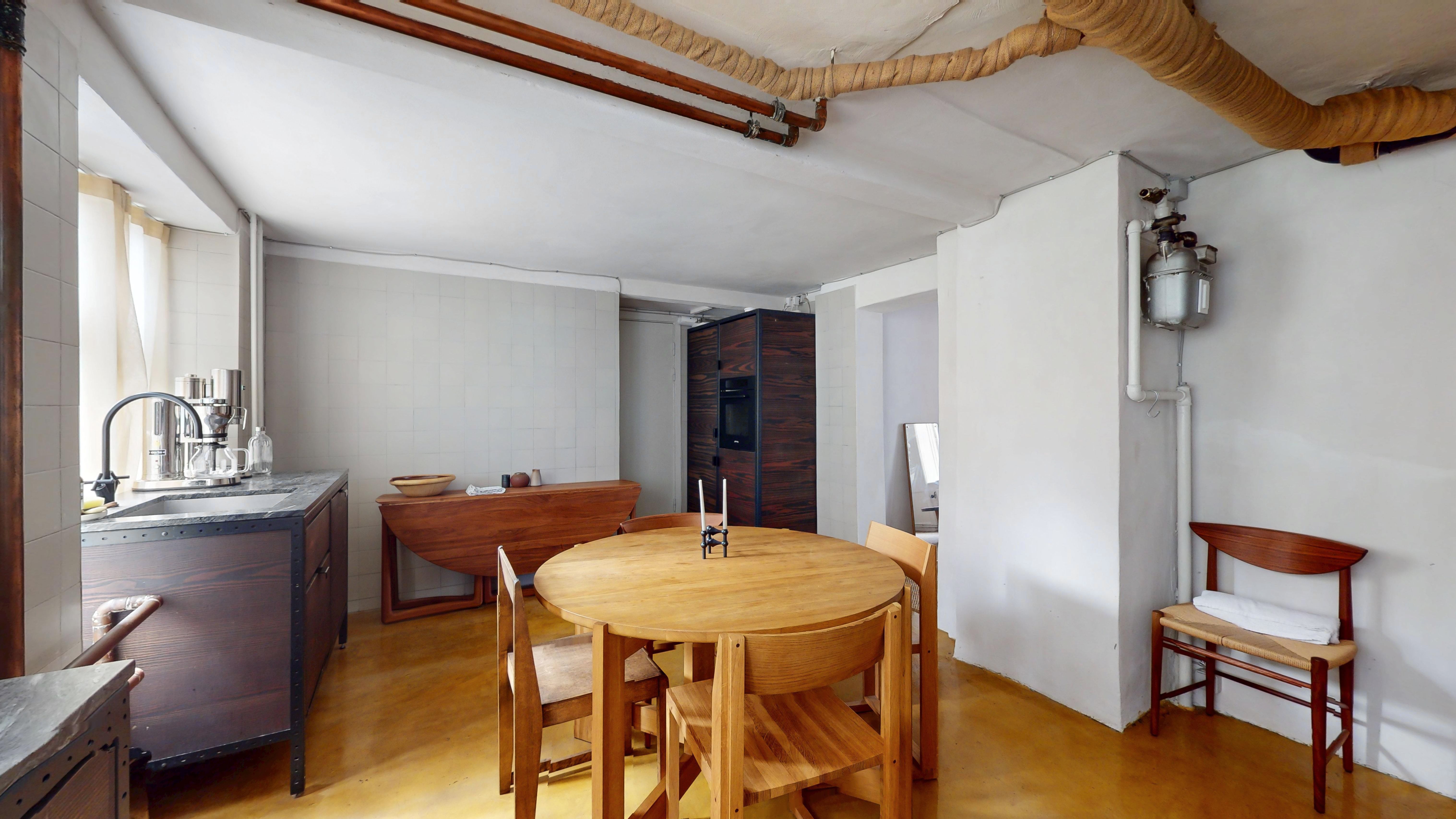 Moderne og minimalistisk stue med lysegul gulvbelægning, runde træbord og stol, rustfri køkkenhøj med vandhaner og et