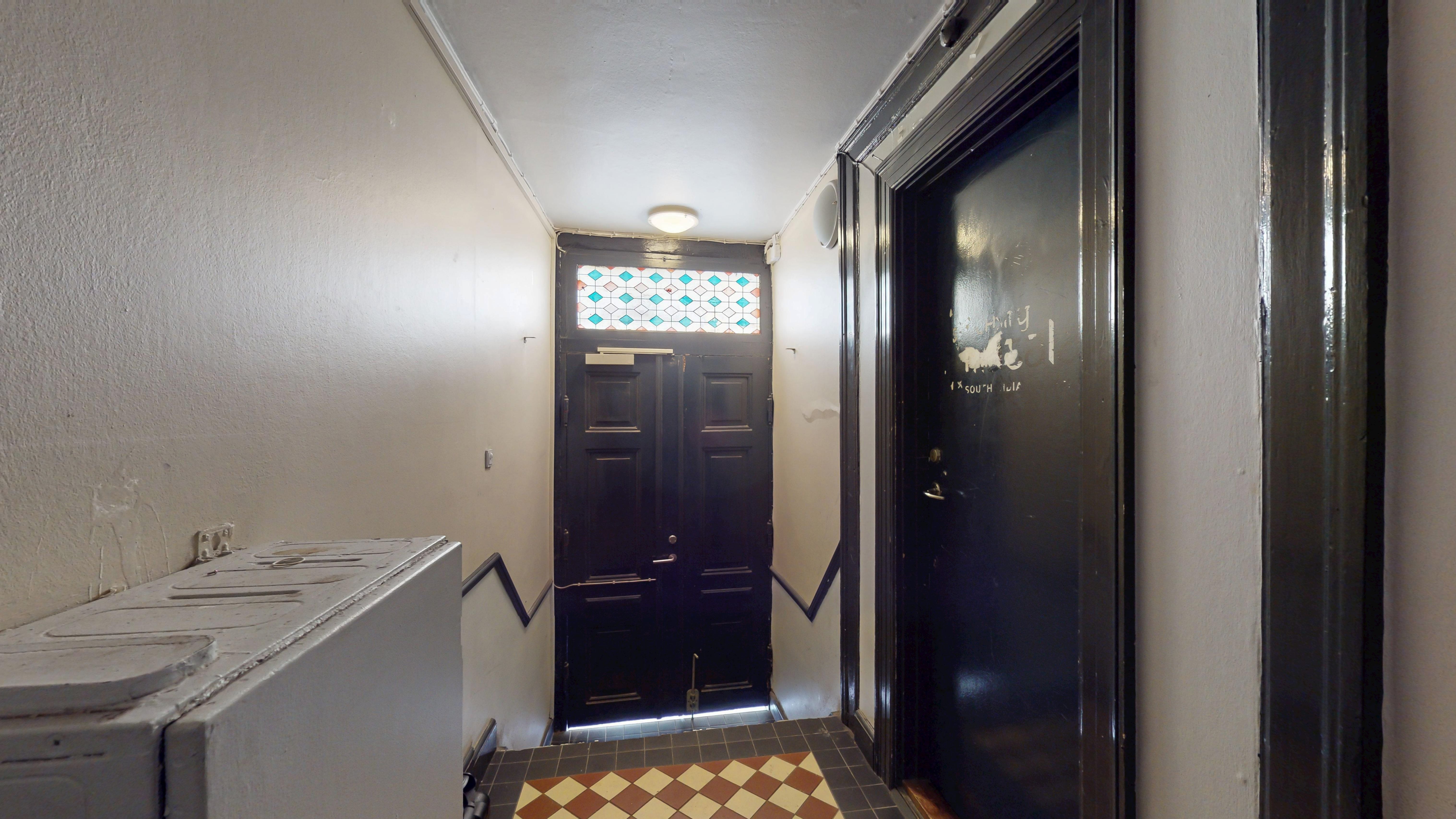 Et langt, tæt og mørkt korridor med to døre, en med en glasvindue og en lysekron, en elektronisk dørslå og gulvet er klædt