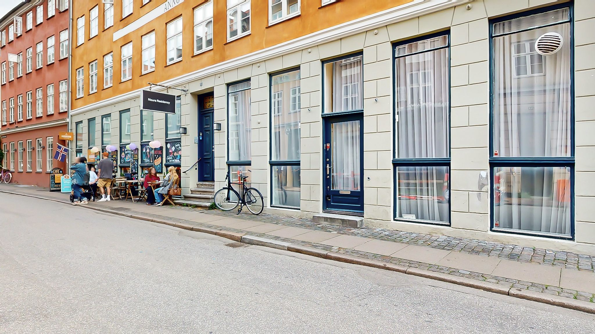 En gruppe mennesker sidder på en terrasse foran en butik, der er indrettet i en rødere og kreativ stil