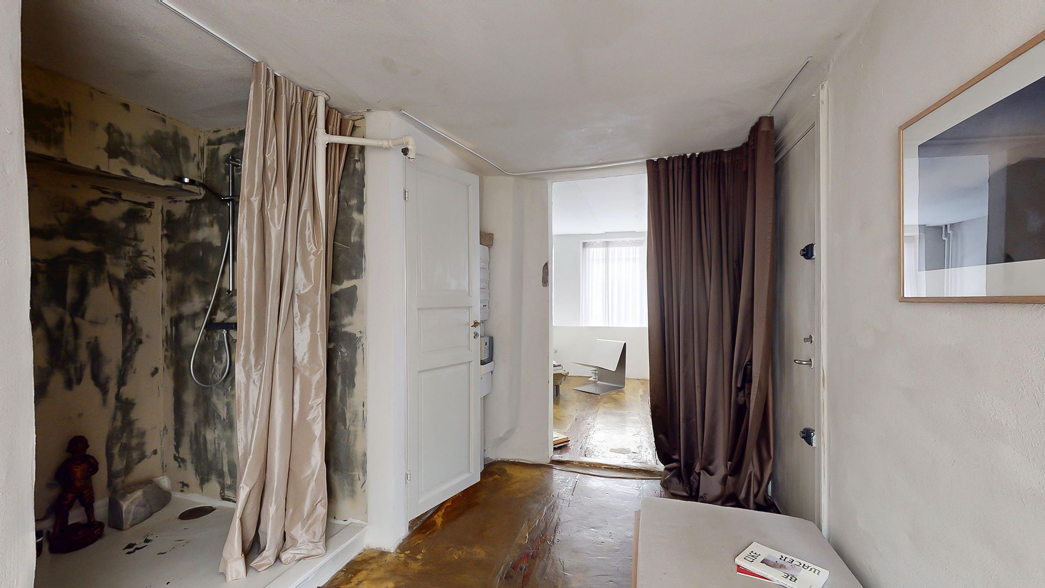 Badrummet er renoveret og har en hvid dør, gulv og loft samt en lukket brusekabine med hængeklæder og et kunstværk på gulvet