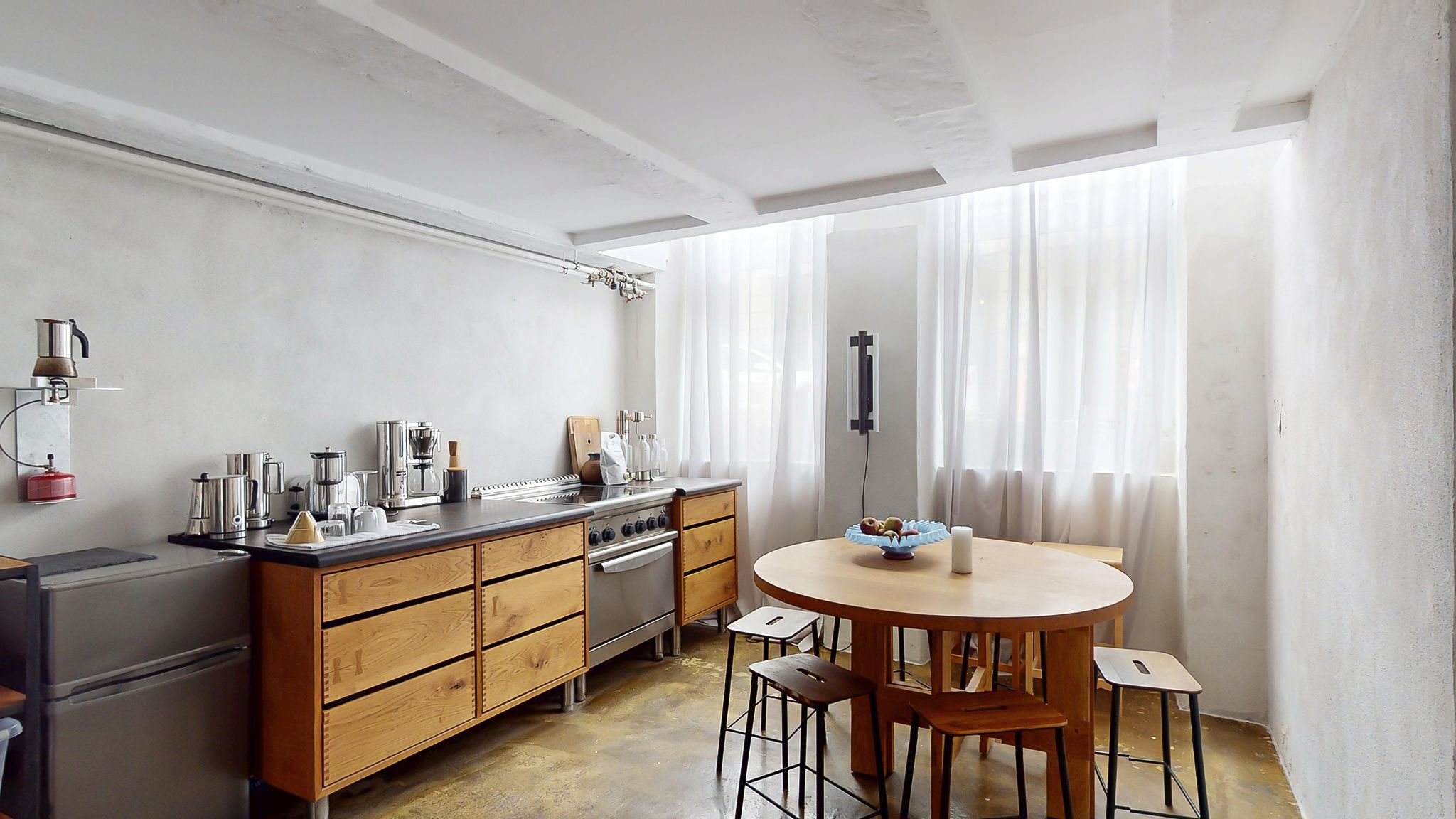 Moderne køkken med højt loft, hvid væg og møbler i bøg og stål. Rundt bord med frugt og en hvid lysestage.