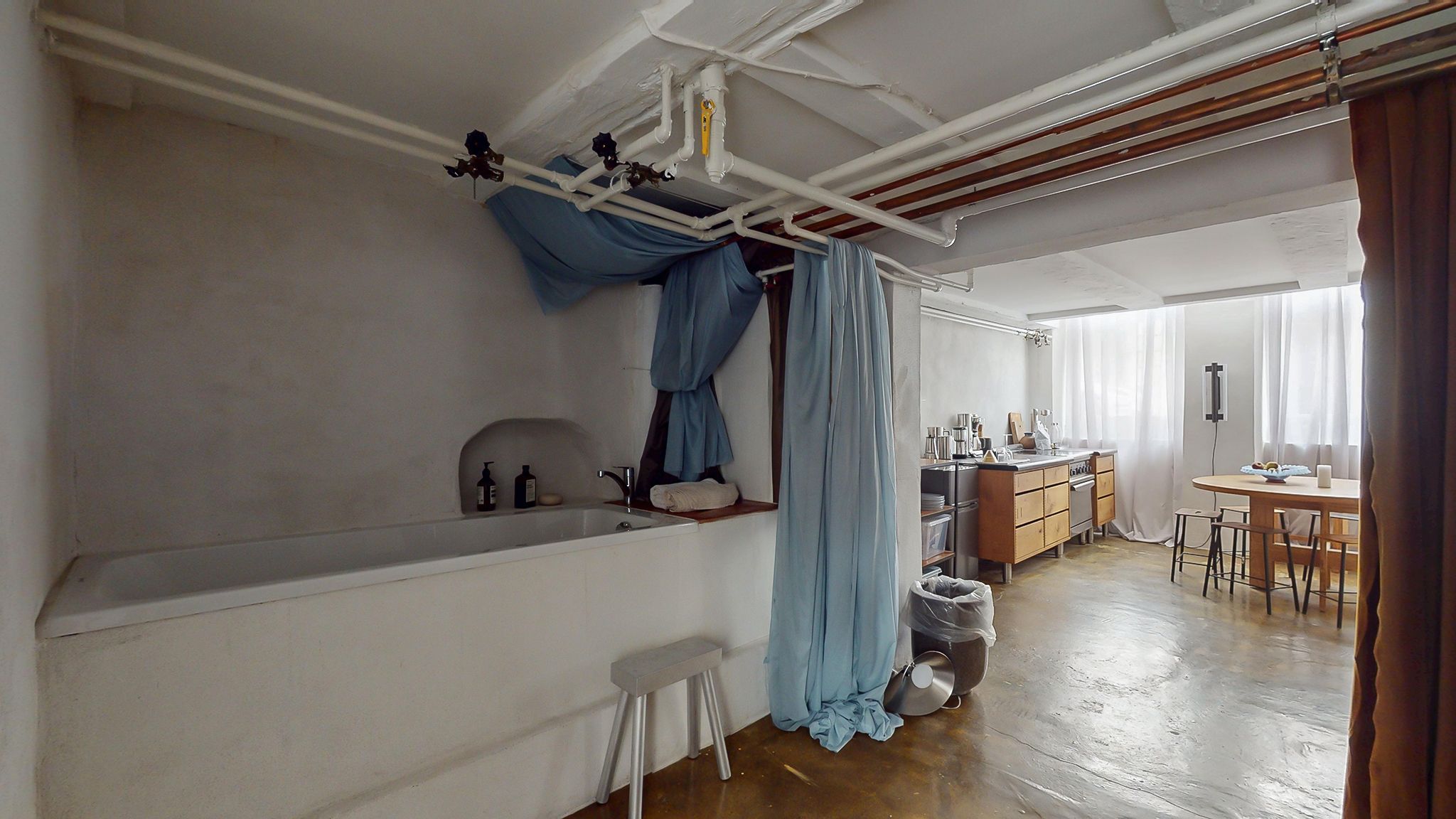 En badeværelse med hvidt badekar og hængende skyggegardiner samt lysegardiner i køkkenet og spisestue med træbord og stole