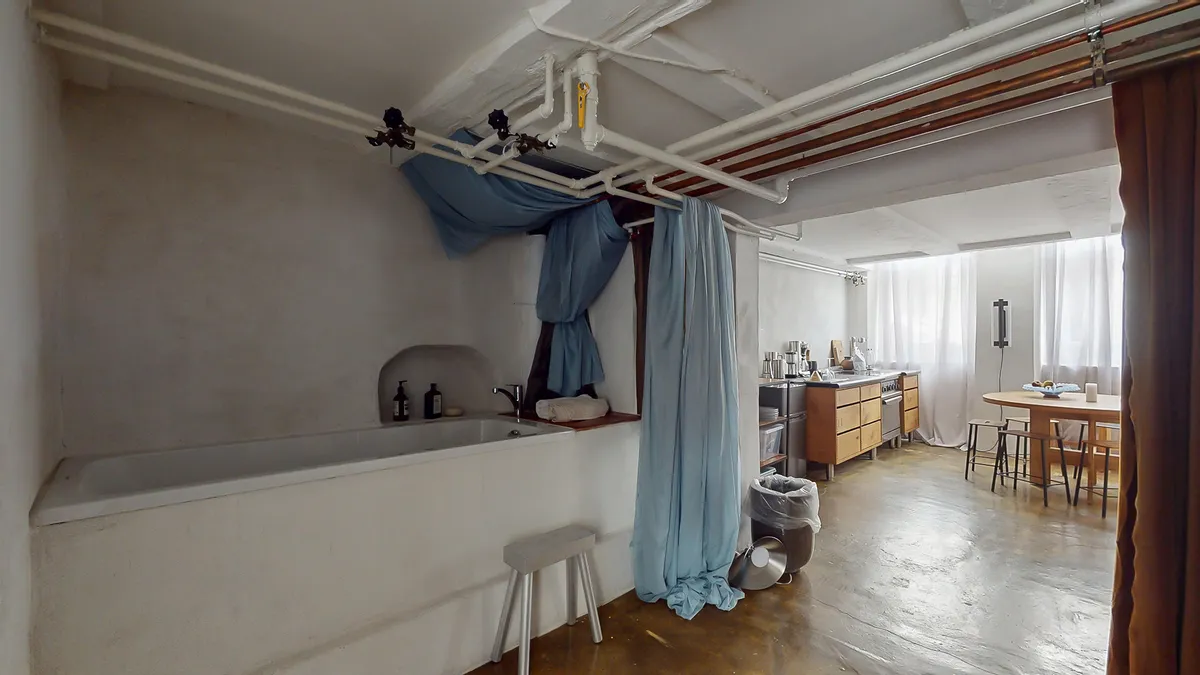 En badeværelse med hvidt badekar og hængende skyggegardiner samt lysegardiner i køkkenet og spisestue med træbord og stole