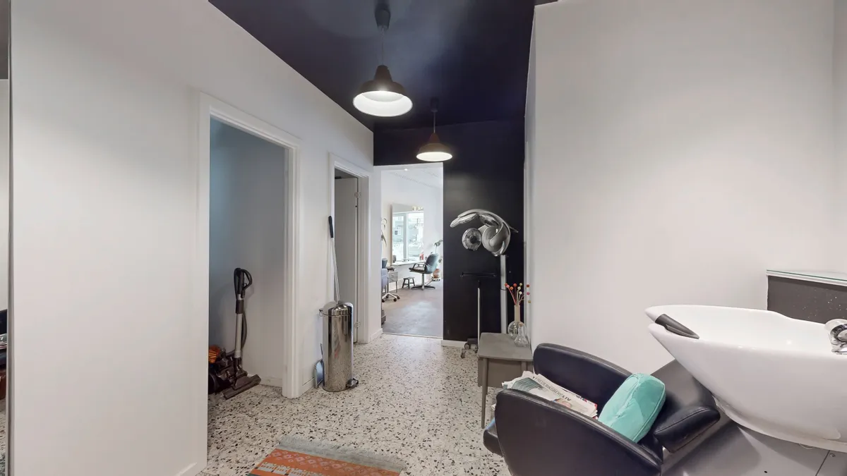 En moderne og pæn frisørsalon med hvid gulvbelægning og mørke lofter samt en stor hvid vaskeskål og en lænestol med pude