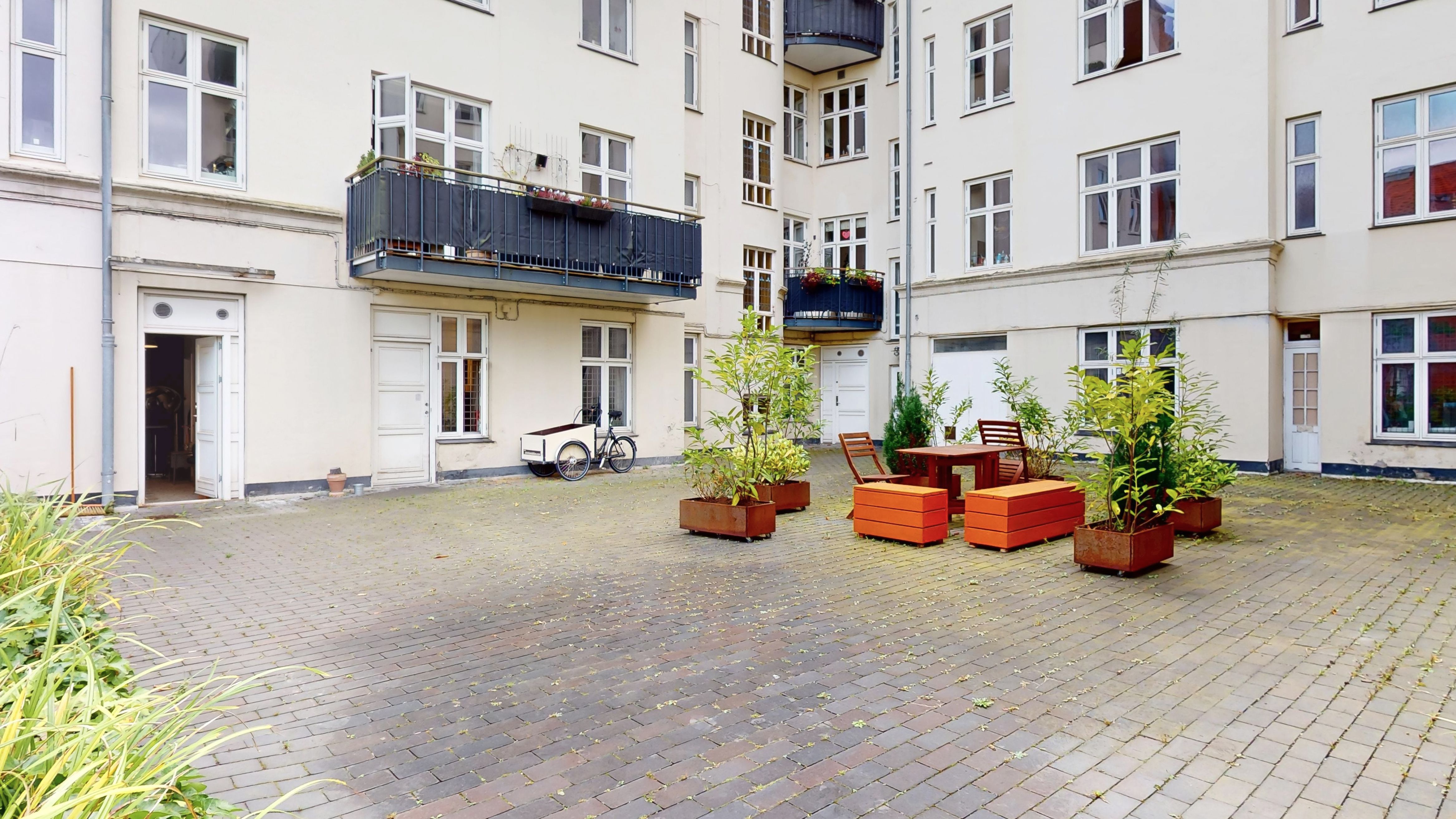 Plantepotter med grønne planter udenfor erhvervslokaler, terrasse med blomster og røde bænke. Både gårdhave og balkon er