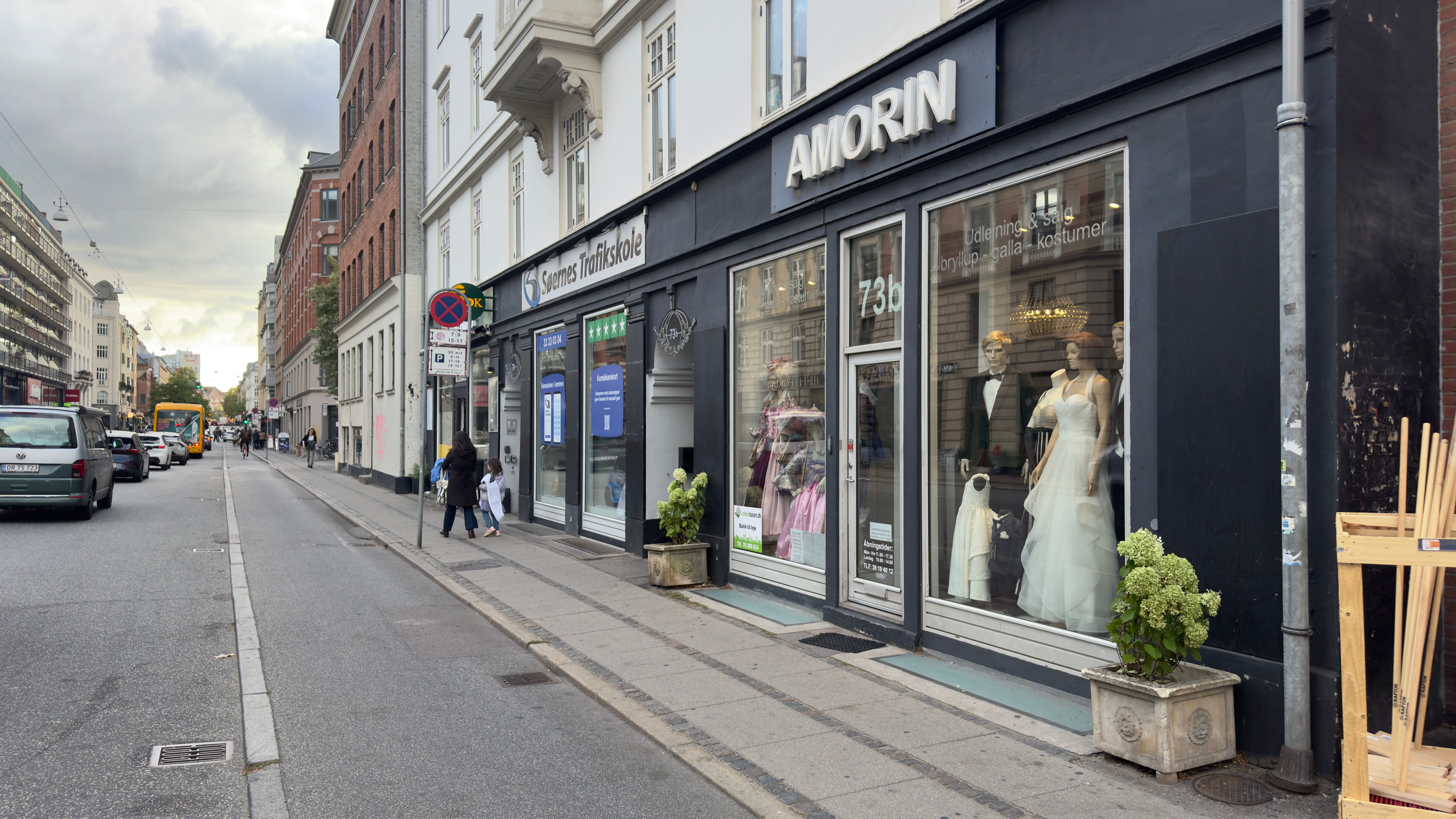 Mennesker går på gågaden foran Amorin butik, der sælger bryllup, gala og kostumer. Der er en bil og en bus i baggrunden.