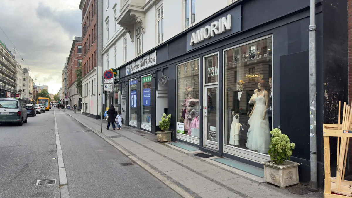 Mennesker går på gågaden foran Amorin butik, der sælger bryllup, gala og kostumer. Der er en bil og en bus i baggrunden.