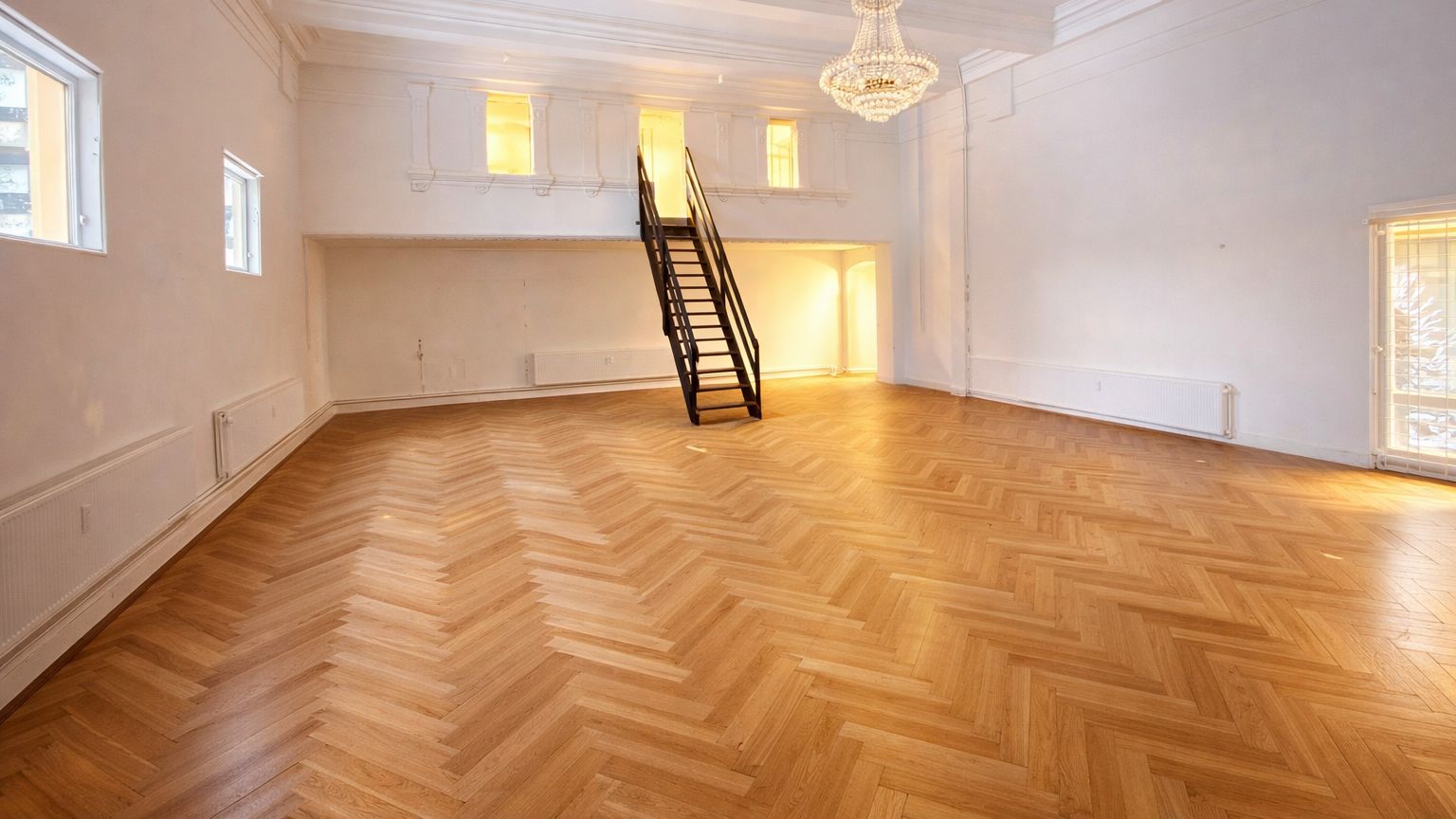 Stort historisk lokale med åbent plan, lyse trægulve og lysekroner. En trappe fører op til første sal og flere vinduer. Ingen mennesker i rummet, der er ren og velplejet