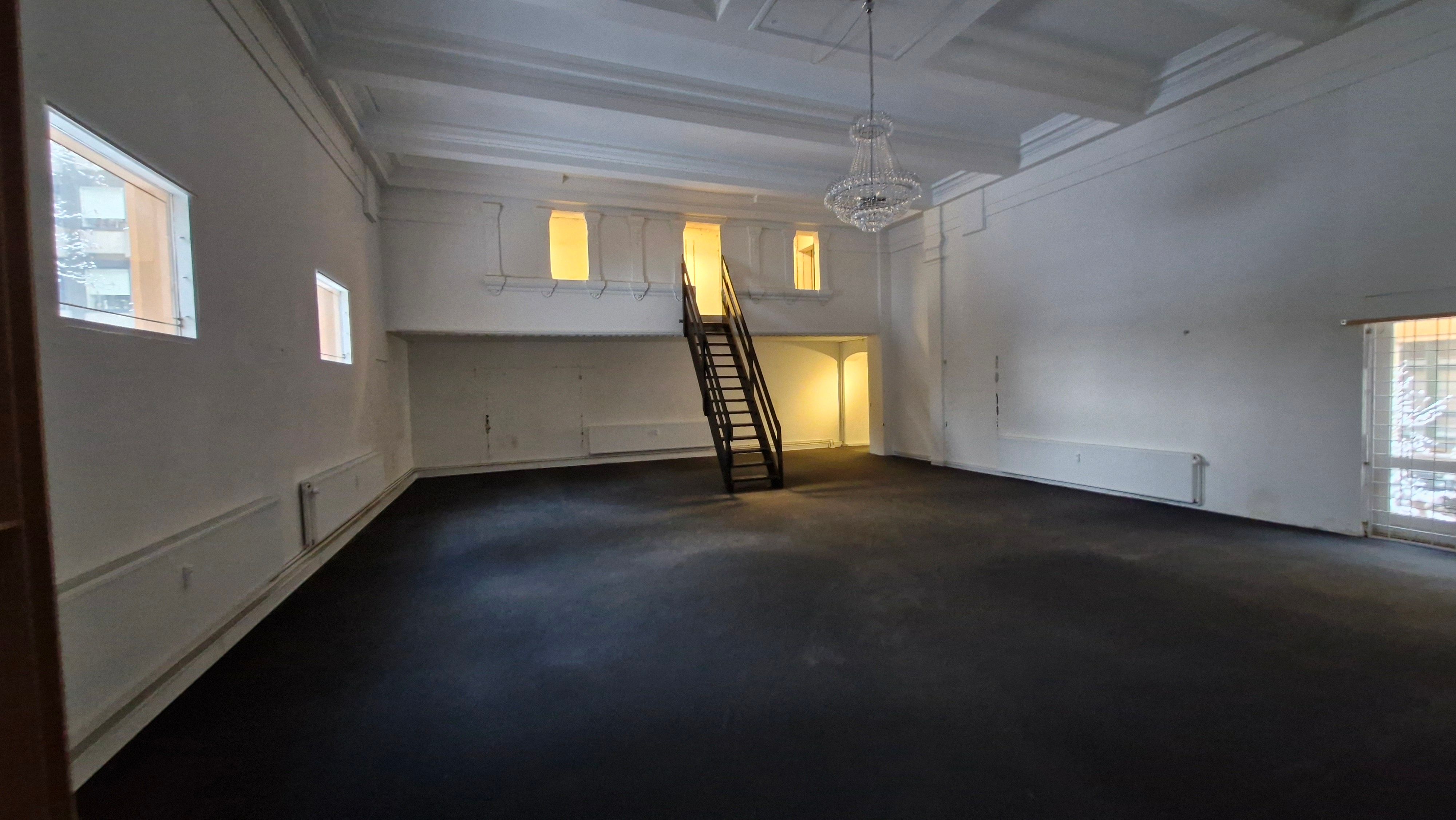 En stor, tom butik med højt til loftet, hvidt vægmaleri og mørke gulve. Stairs i midten fører op til første sal. Lys kommer ind gennem vinduer og et stort krystalkronelys.