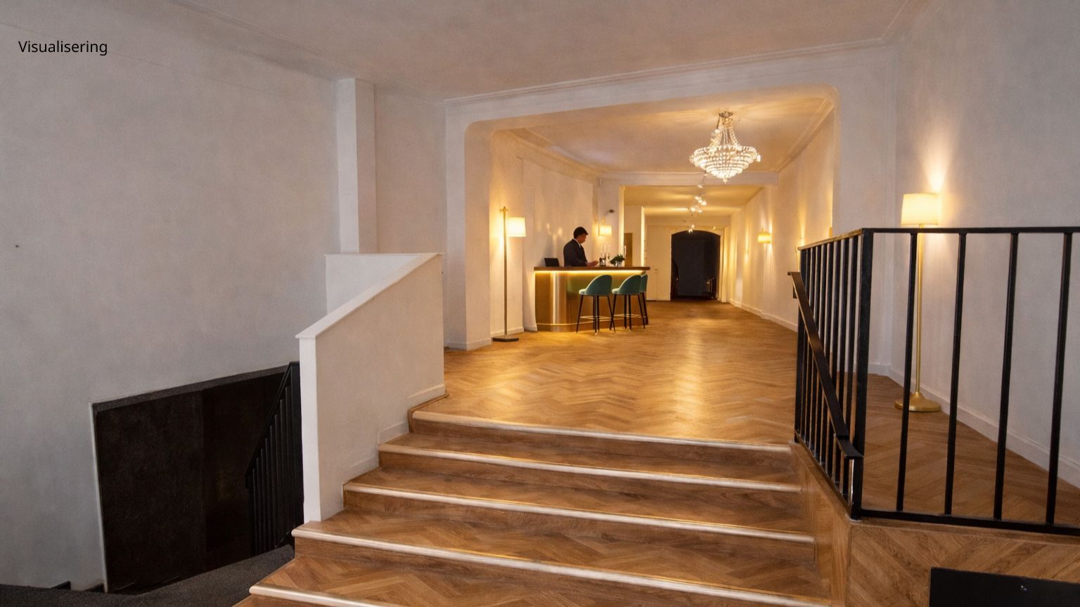 Elegante trægulve og hvide vægge. En mand i sort sidder ved et bord med en computer. To grønne stole med guldfarvede ben står ved bordet. Lyskroner og lysestager hænger fra loftet.