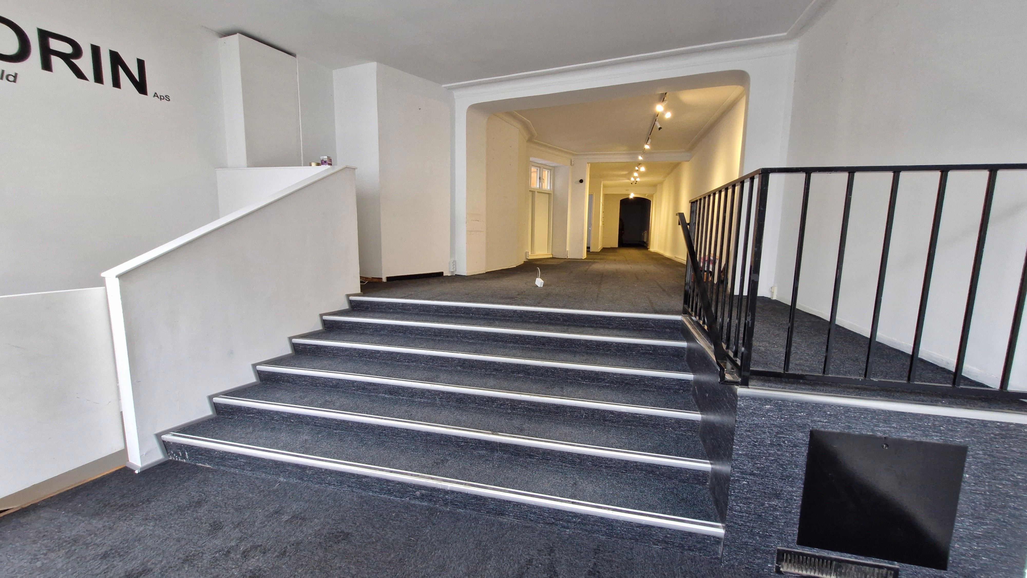 Styretrappet med mørke trin og lyse kantstriper fører op til indgangen af et erhvervslejemål. Væggen har ordet Dorin i mørtegn og lyse punktlys i loftet