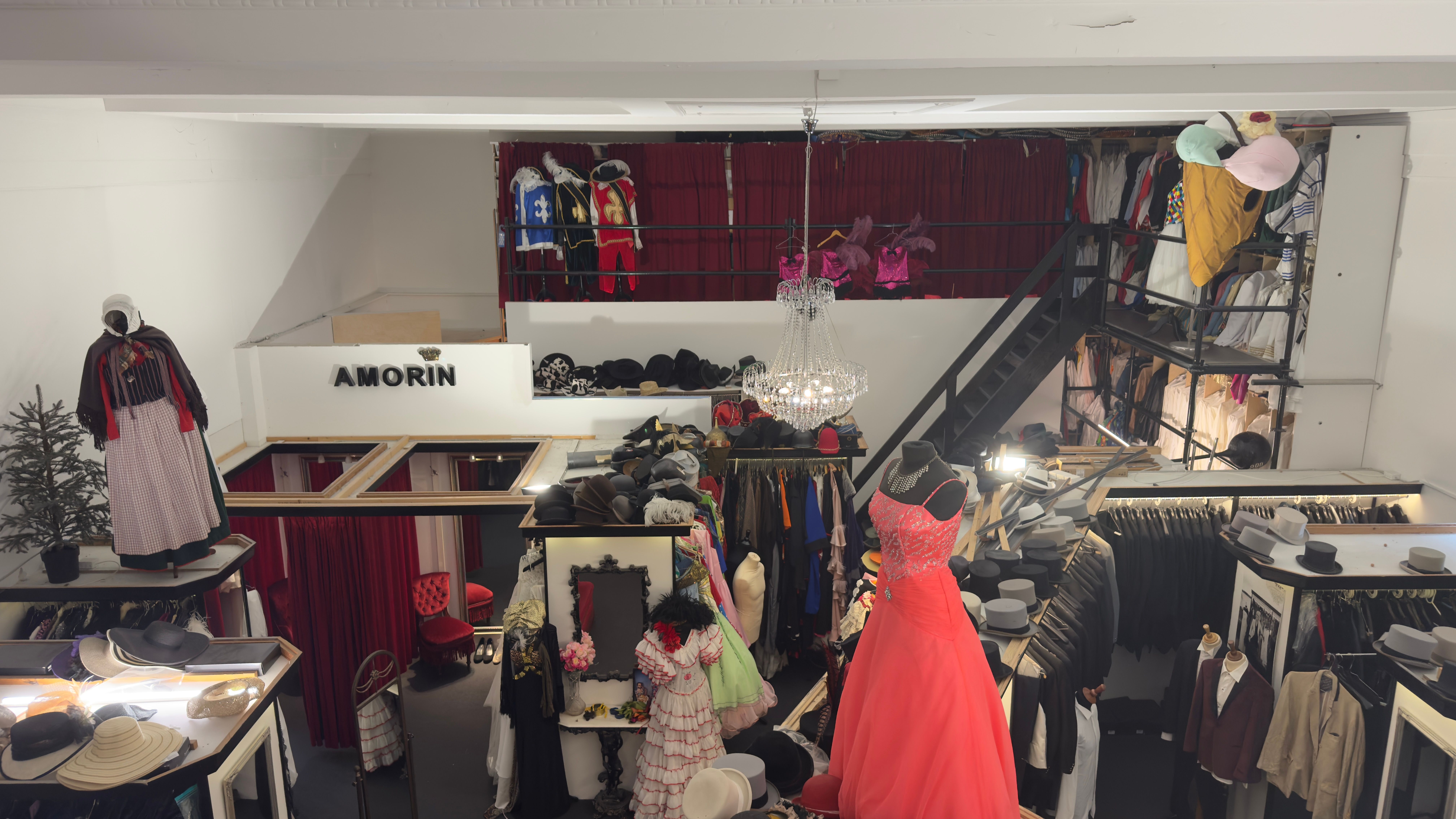 Forretningslokal med mange kjoler, hatte, kostumer og andet. Kjoler og kostumer hængende på klædstativer. Hatte på hylder.