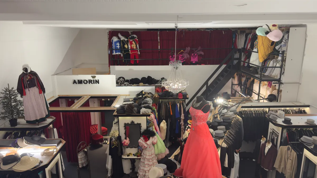 Forretningslokal med mange kjoler, hatte, kostumer og andet. Kjoler og kostumer hængende på klædstativer. Hatte på hylder.