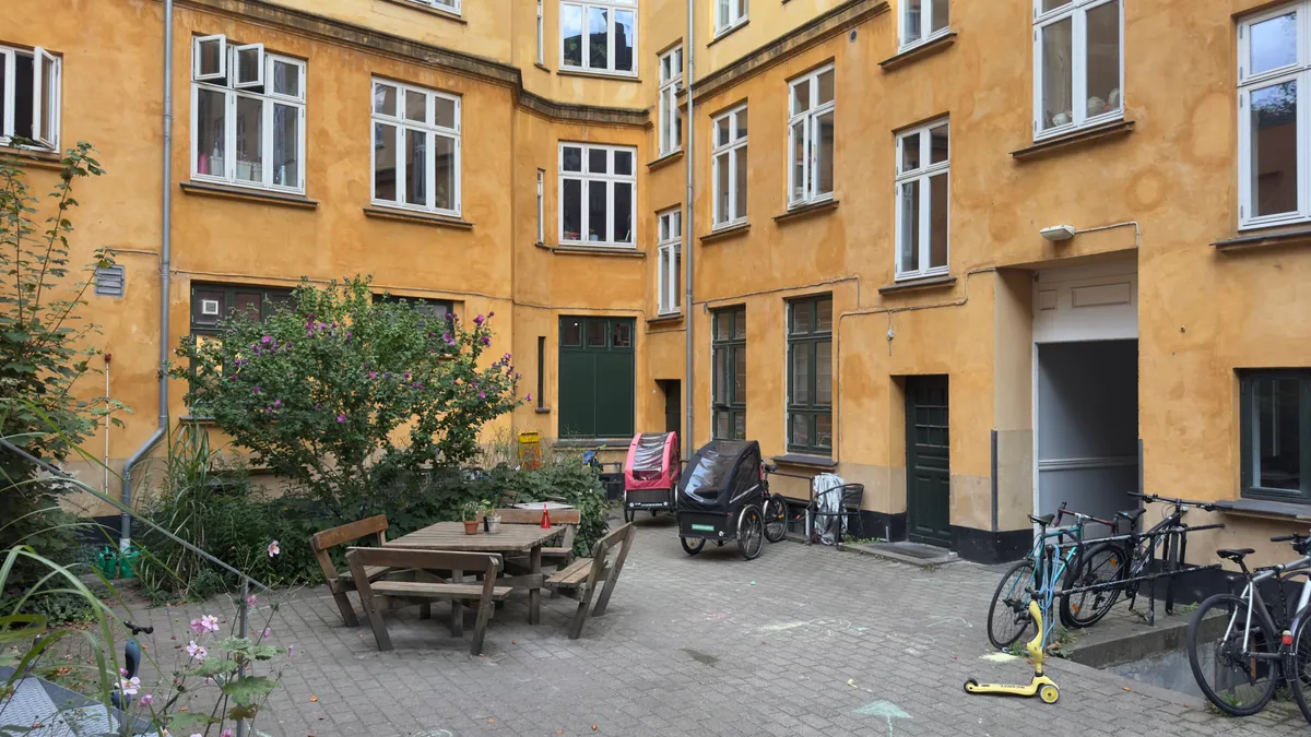 En smuk gårdhave med træbord og stole, to trækvognstrailere, fire cykler og en trækvogn i bakke op mod en gård.