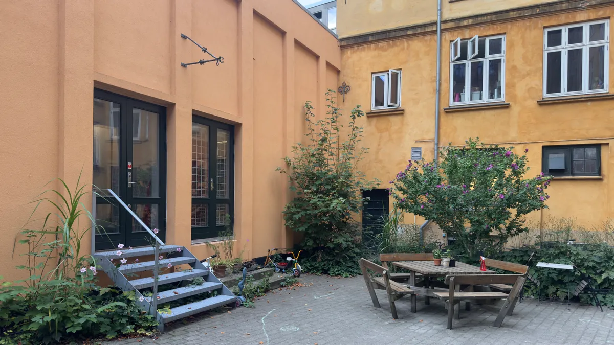 Smuk gårdhave med træbord og stole, trætrapper, grønne planter og blomster, gul bygning med vinduer og døre