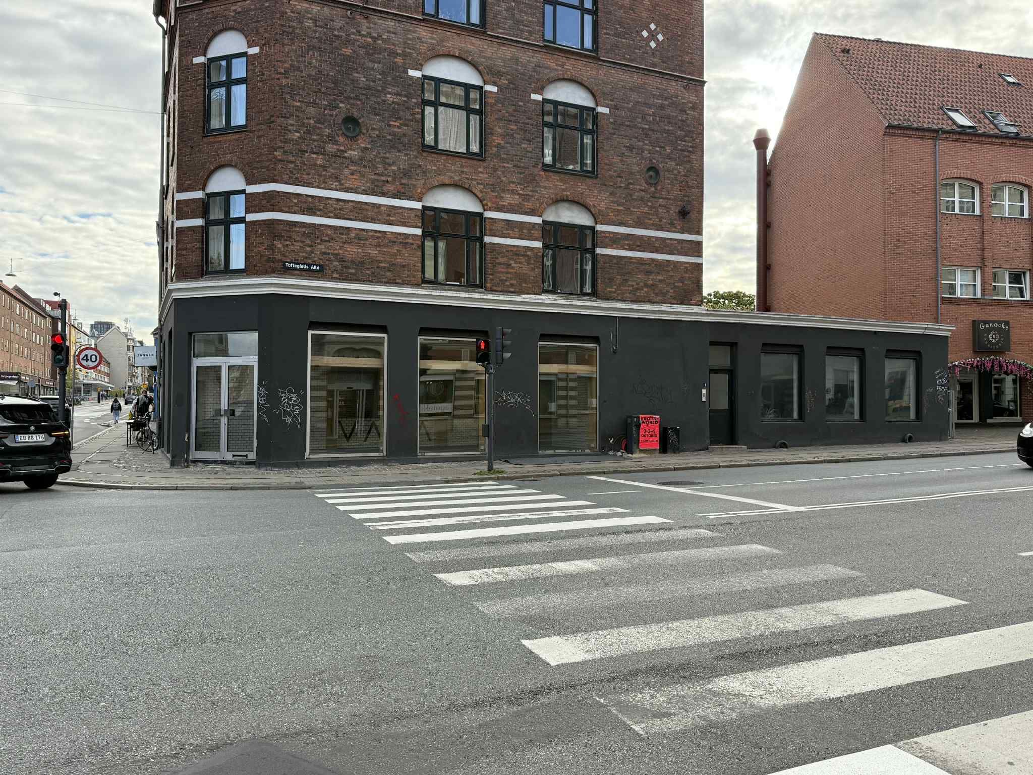 Gade med tomt parkeringsfelt og kryds. Adgangsvej til et erhvervslokal med glasruder og trafiklys. Hjørnebygning med røde