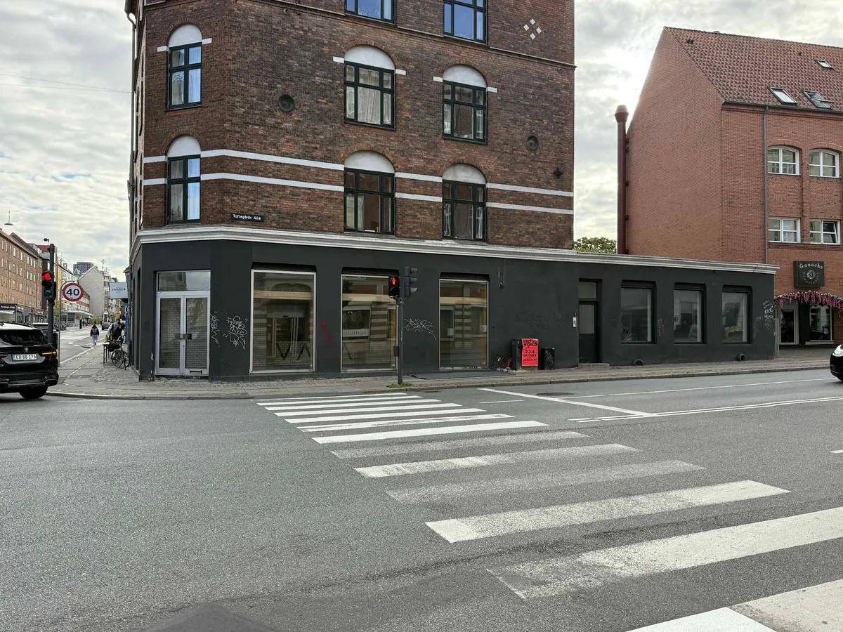 Gade med tomt parkeringsfelt og kryds. Adgangsvej til et erhvervslokal med glasruder og trafiklys. Hjørnebygning med røde