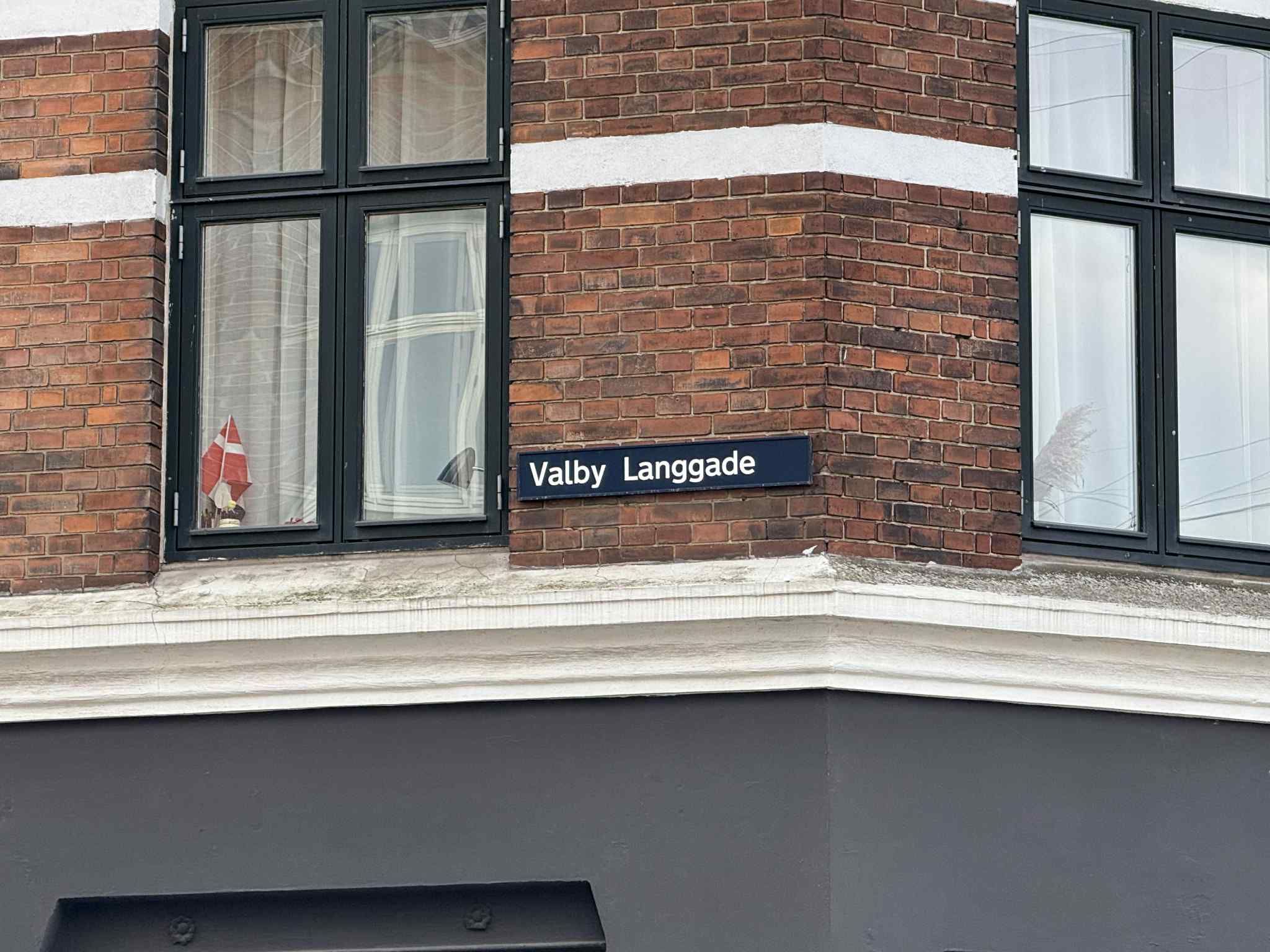 En teglbygning med vinduesglas og et navneskilt på Valby Langgade, der tyder på en bygningsadresse.