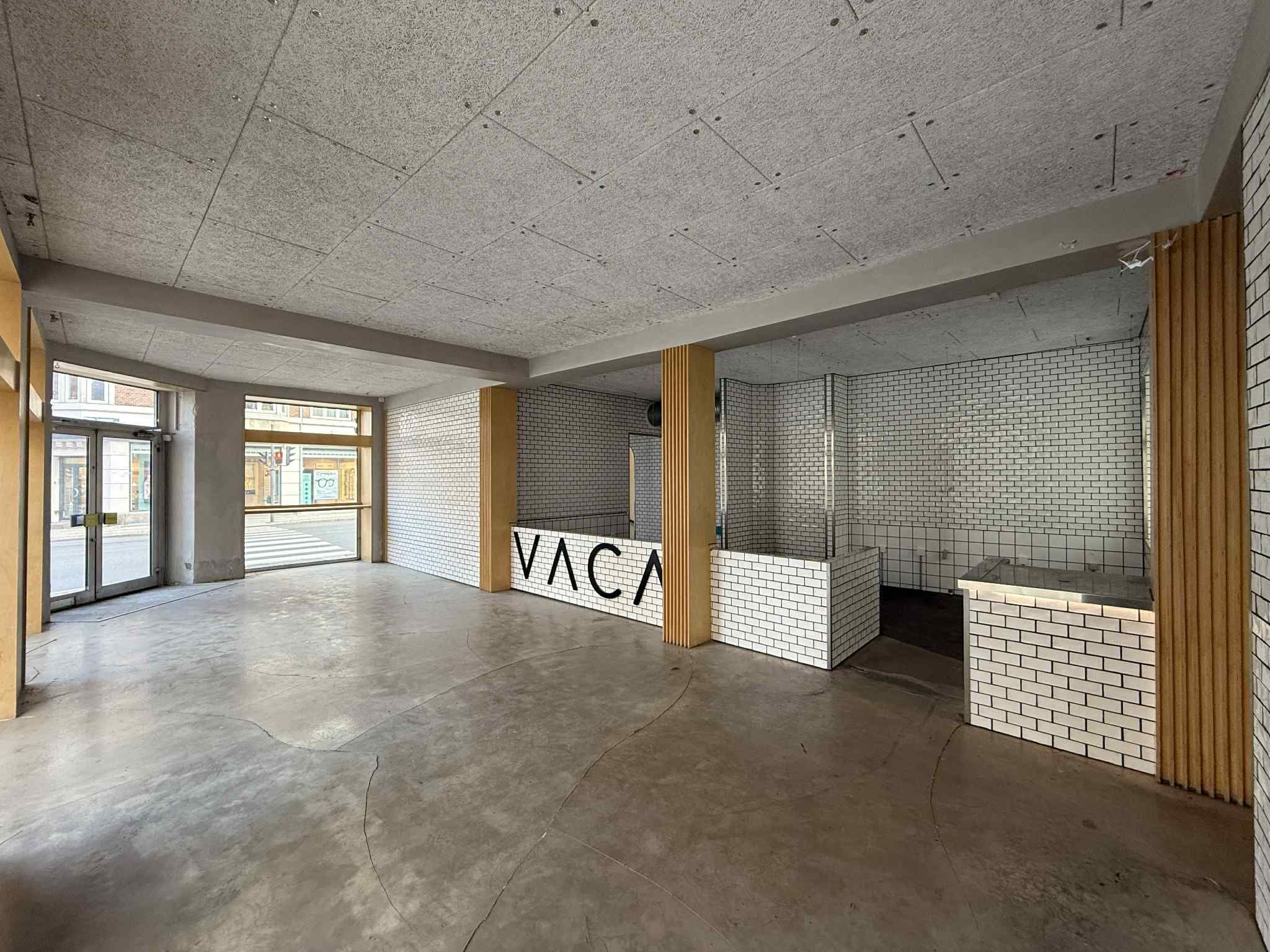 Tom lokale med glasdøre og vinduer, højt loft, hvidt tøj, møbler og teksturer, og "VACA"-logo på væggen