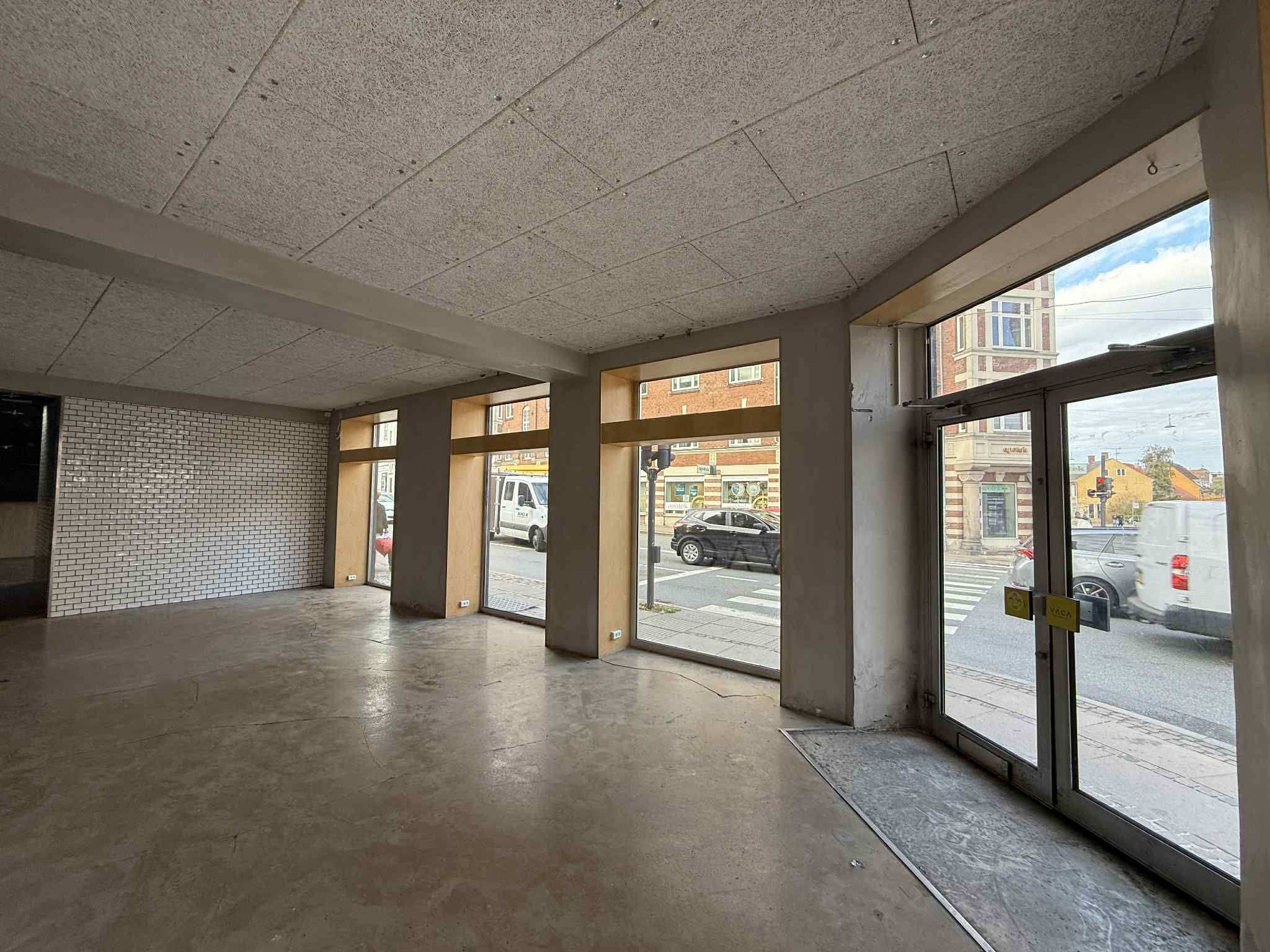En tom erhvervslokal med højt loft og lyse vinduer og store glasdøre med udsigt til gaden med biler og bygninger