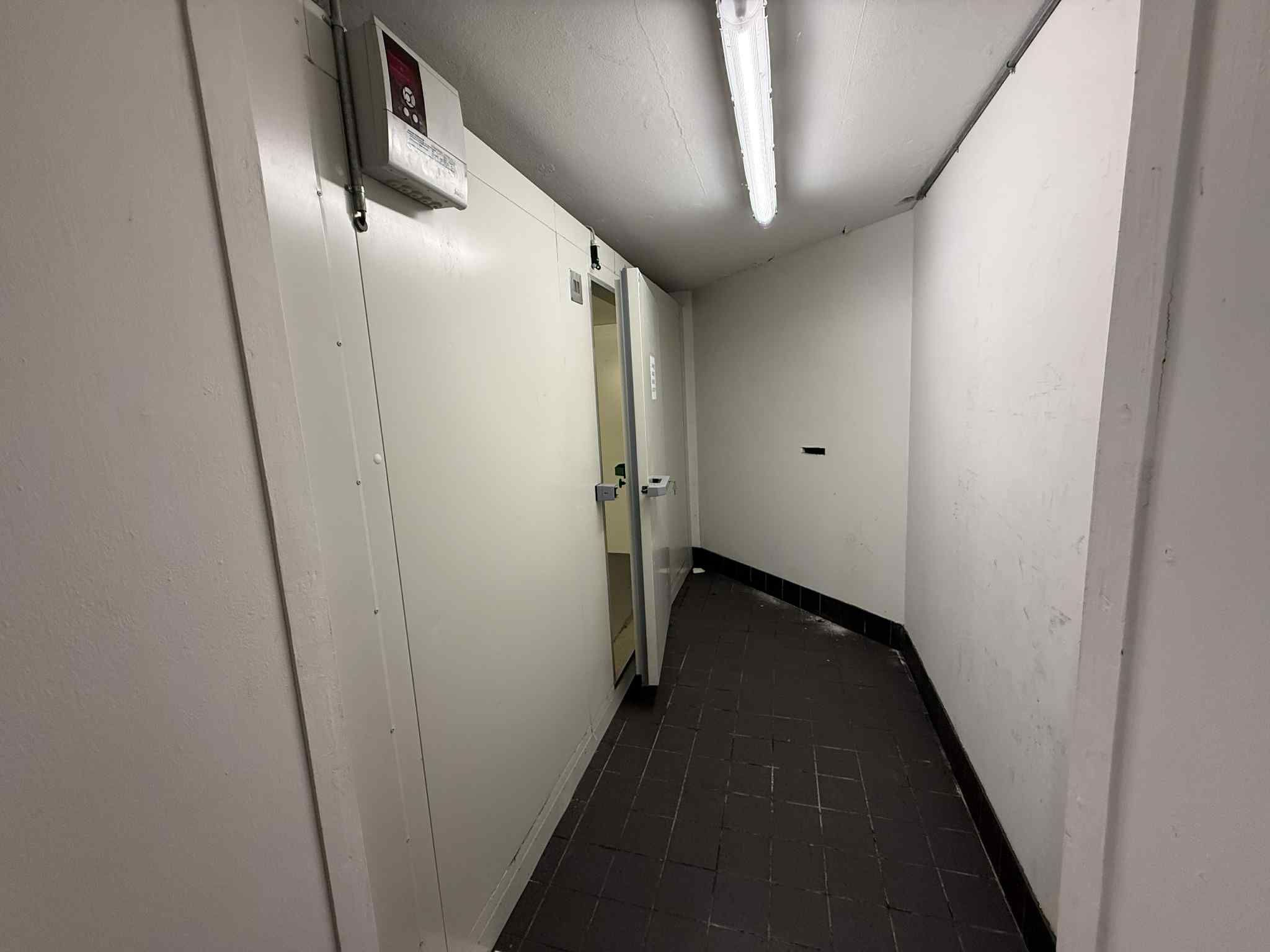 En kølighusetgang med lyse hjørnevægge og gulvtæppe, glasdør og en betjeningsstation med en tændt lampe