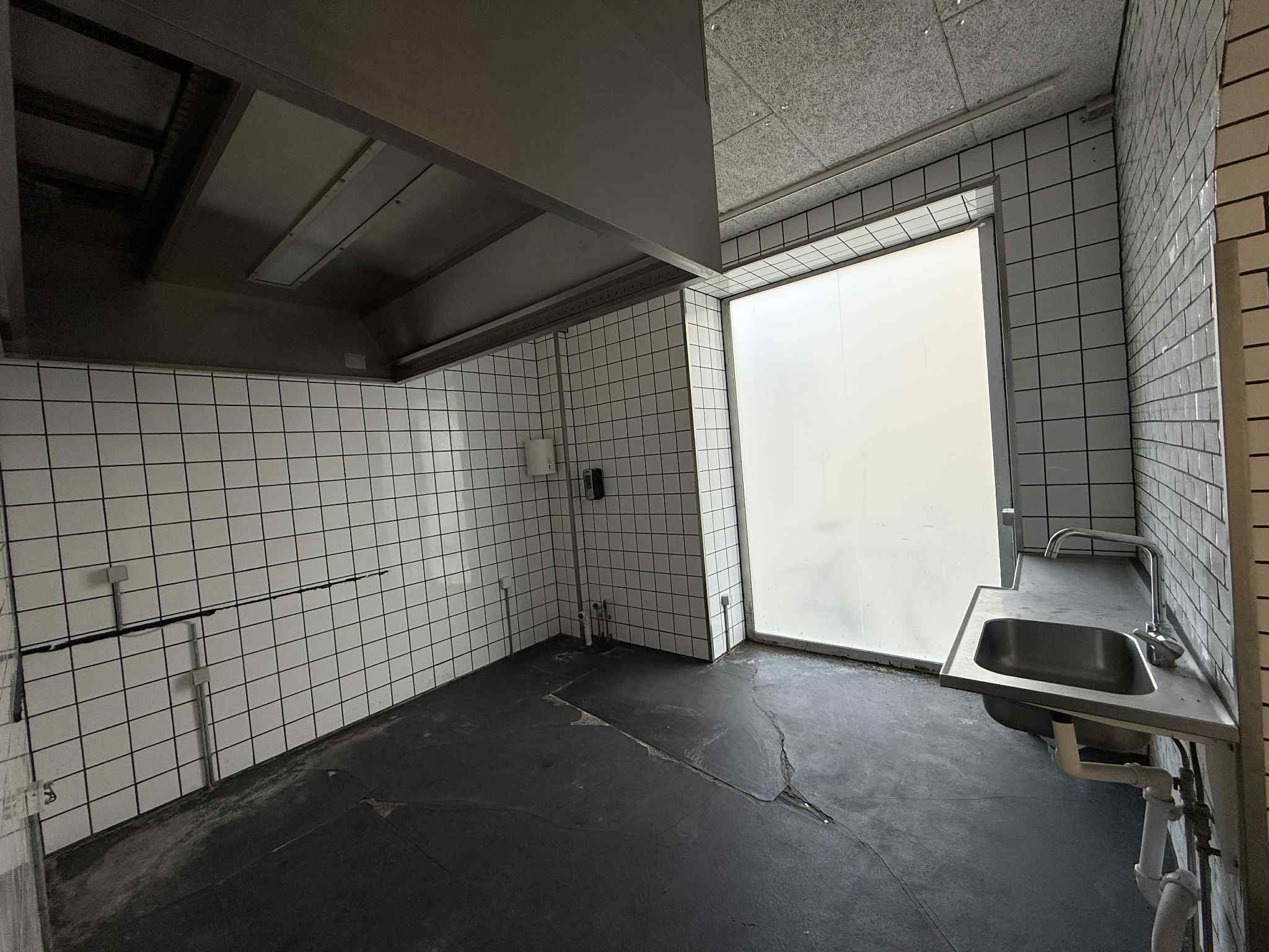 En tom værkstedslokal med sort gulv og hvide flisede vægge, en metalvandhane og et håndvask med en lukket dør til en