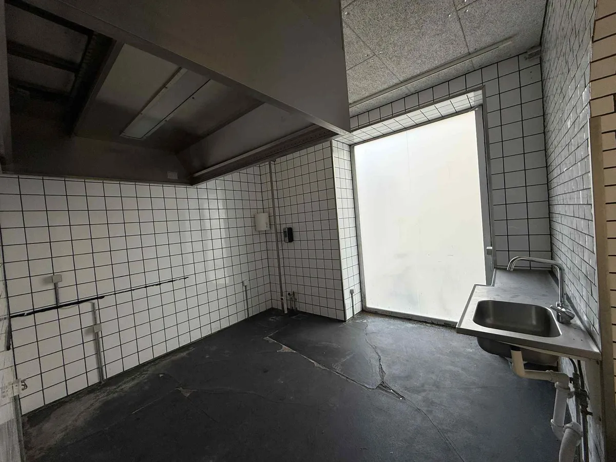 En tom værkstedslokal med sort gulv og hvide flisede vægge, en metalvandhane og et håndvask med en lukket dør til en