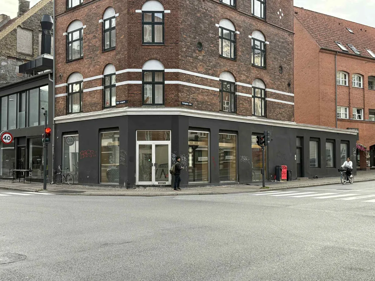 En hjørnebygning med store glasruder og graffiti og to cyklister passerer på en tom gade