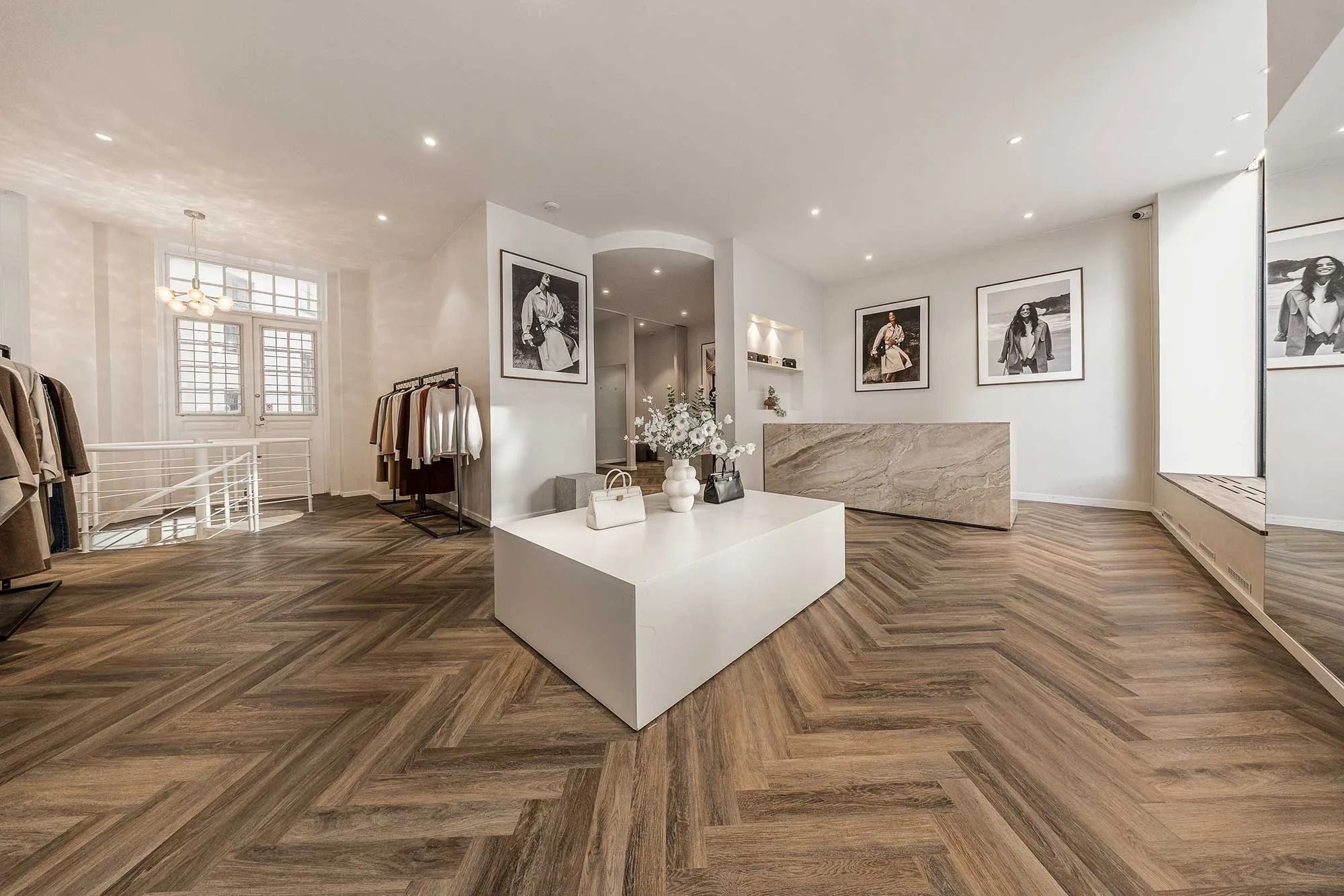 Elegant og minimalistisk butiksindretning med trægulv, hvide vægge og lyse loftslamper. Her er et hvidt bord med blomster og