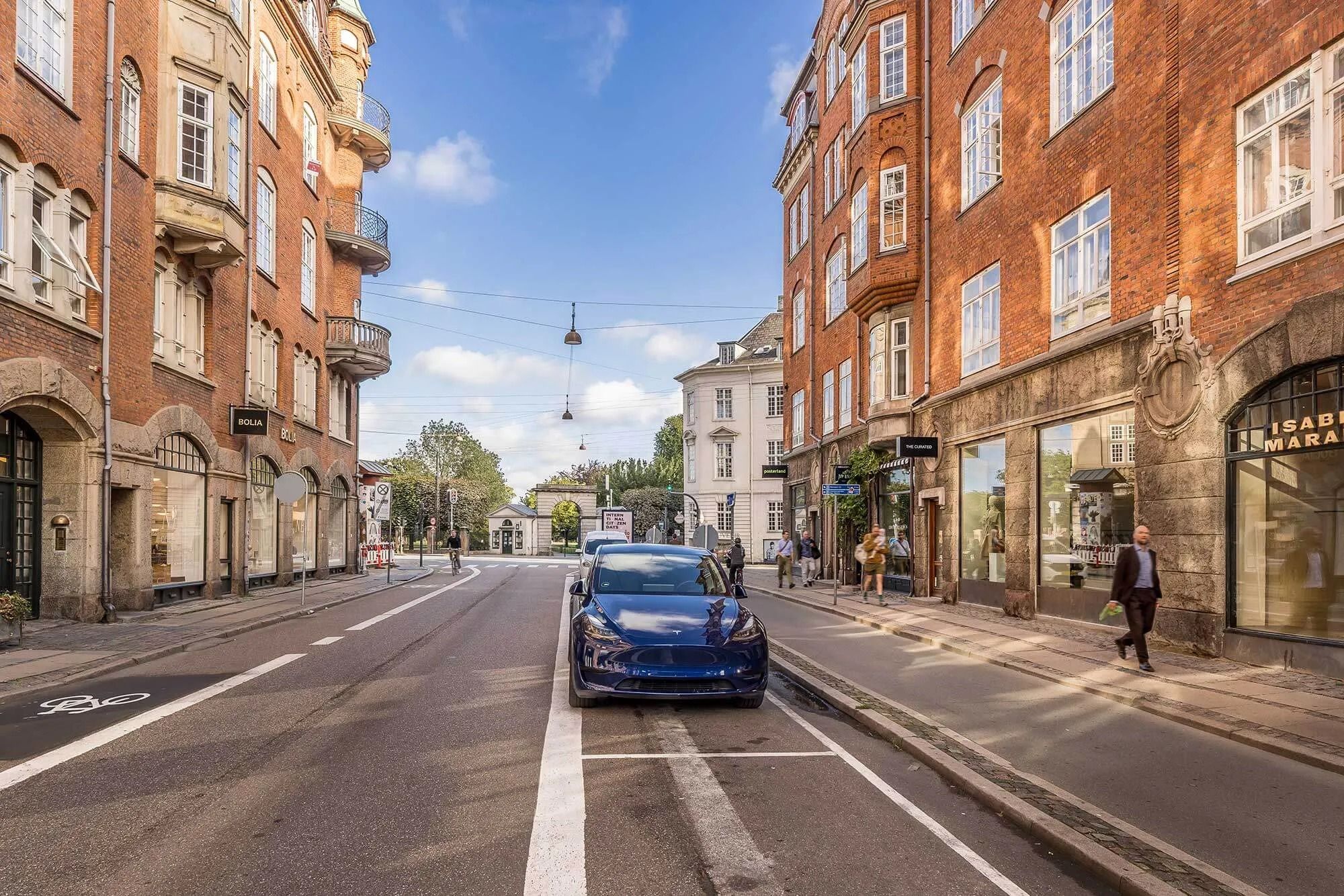 En ny blaue Tesla Model 3 kører gennem en gågade i København med butikker og huse med facader i rød mursten