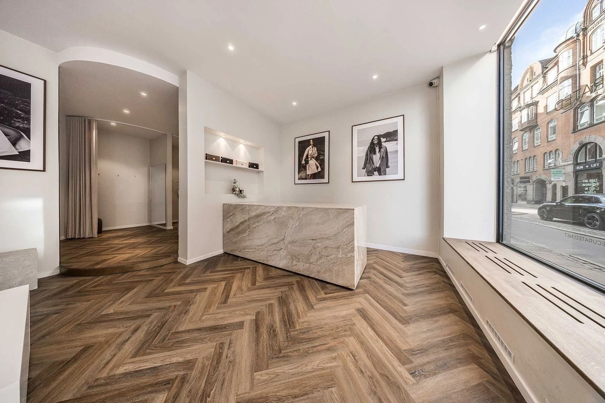 Flot og elegant interiør med hvidt loft, marmorbord og parketgulv i en butik. Vindue med udsigt til en bil og bygning. Væg