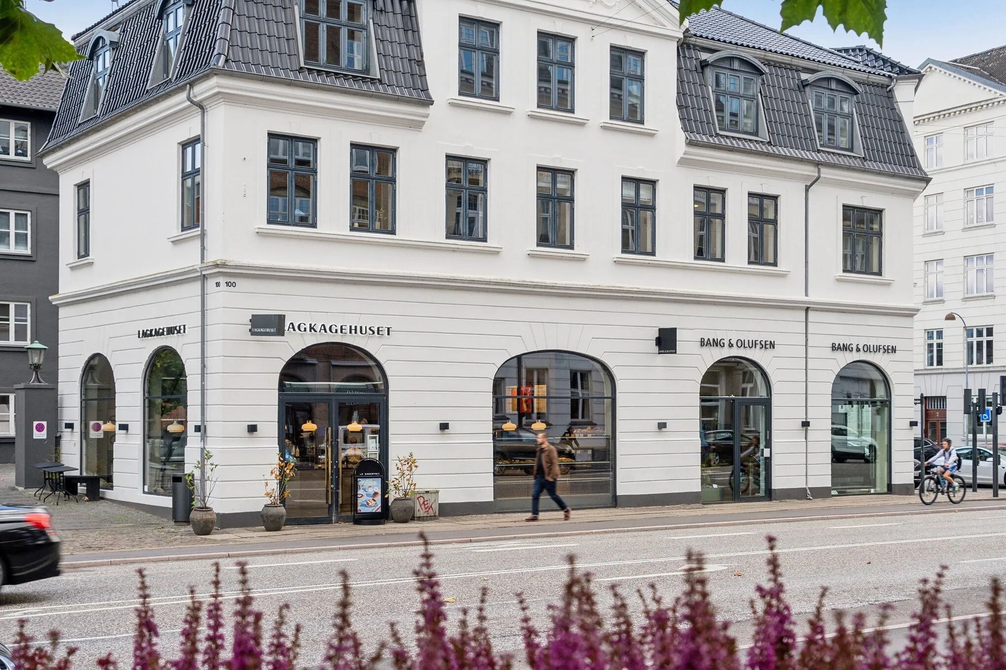 Man går forbi en hvid bygning med Bang & Olufsen butik og AKKAGEHUSET logo, der er dekoreret med plantepotter og lampe