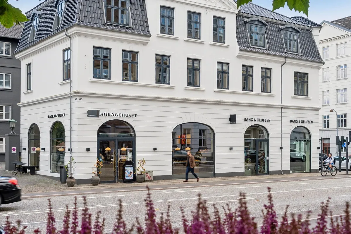 Man går forbi en hvid bygning med Bang & Olufsen butik og AKKAGEHUSET logo, der er dekoreret med plantepotter og lampe