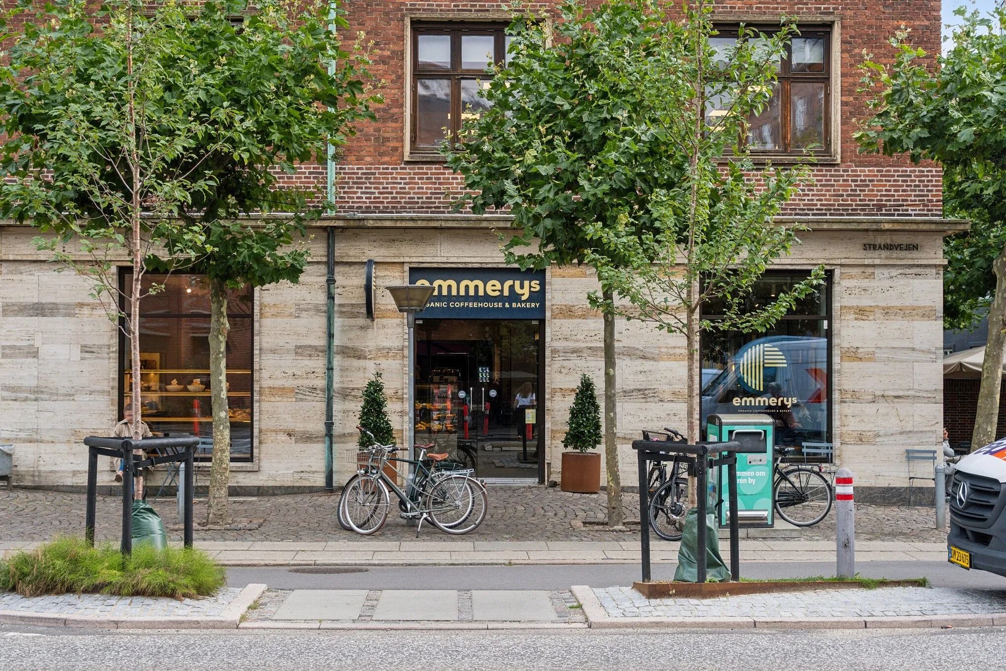 Cykler står parkeret udenfor Emmerys café og kagebakeri med udsigt til flere træer og blomsterpotte