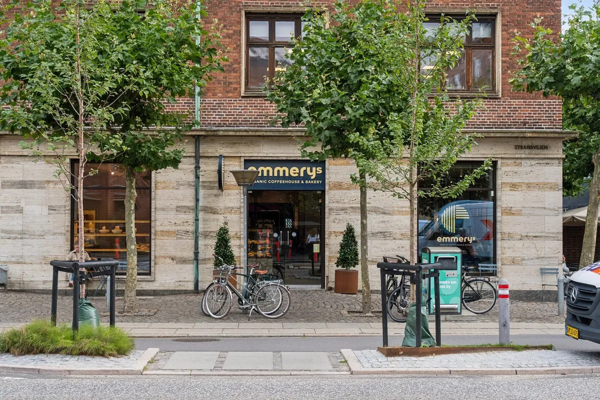 Cykler står parkeret udenfor Emmerys café og kagebakeri med udsigt til flere træer og blomsterpotte