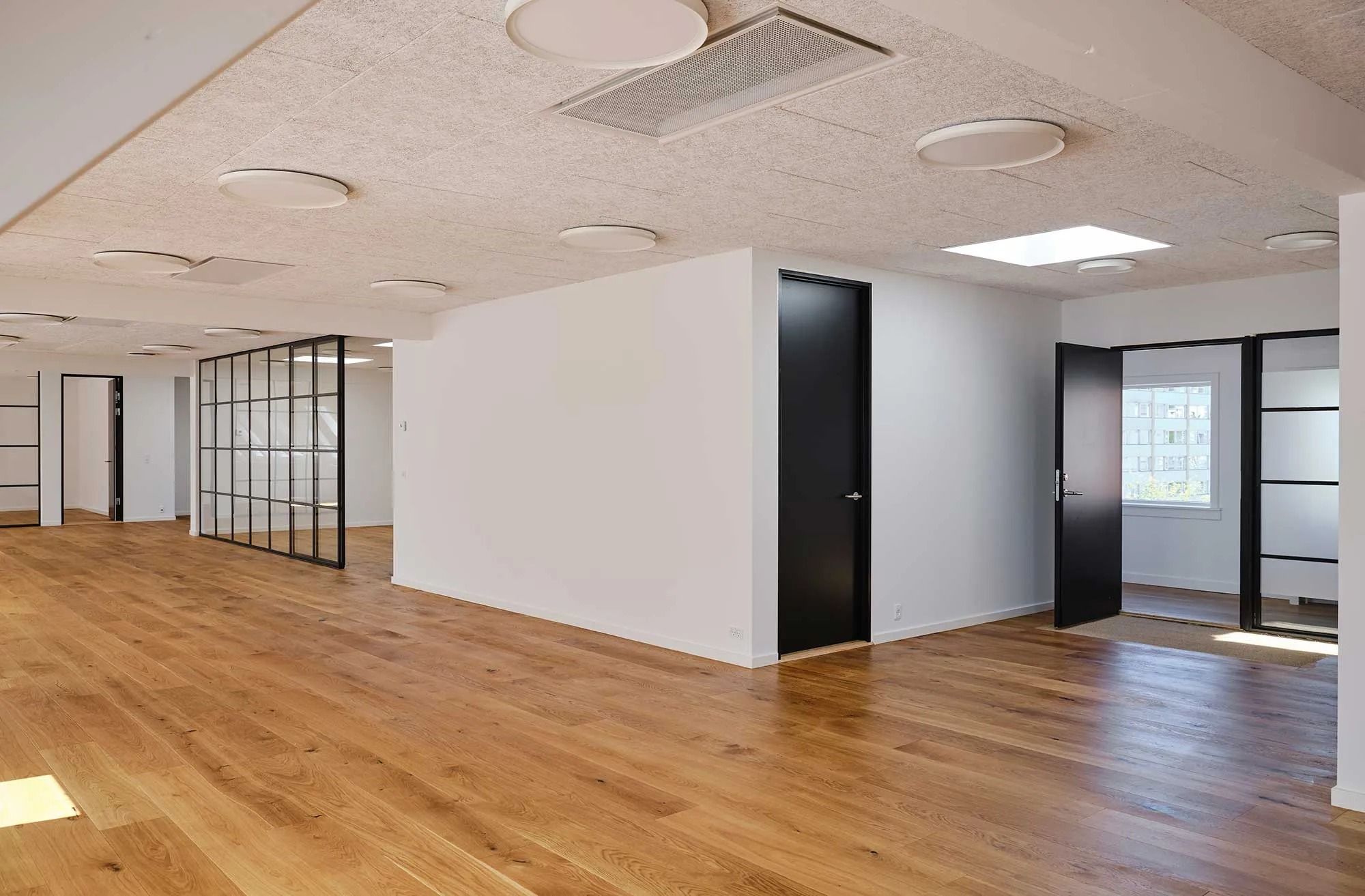 Kontorrum med hvide vægge, lyse trægulve og moderne møbler samt store vinduer med udsigt til byen