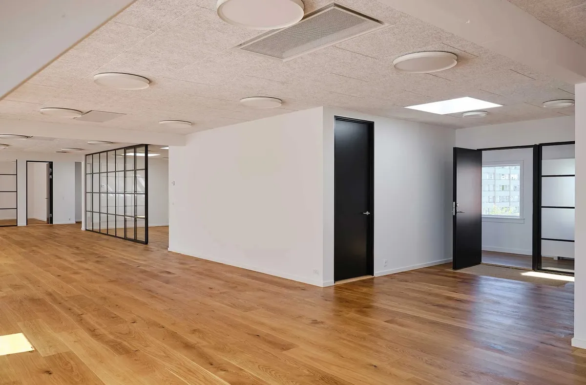 Kontorrum med hvide vægge, lyse trægulve og moderne møbler samt store vinduer med udsigt til byen