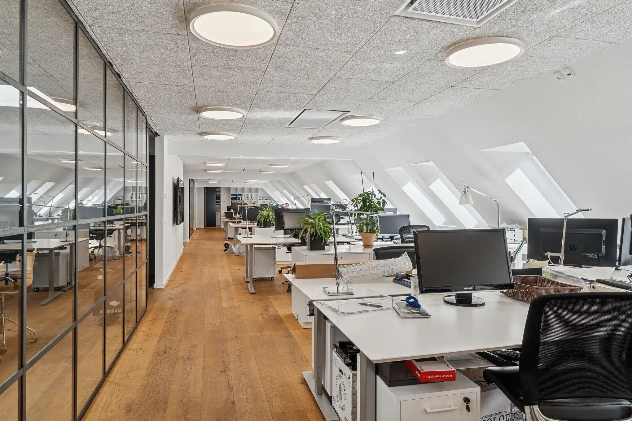 Kontor med højloft og gul trægulv, hvor flere skriveborde og computere er placeret langs væggen og midten af rummet. Rundt