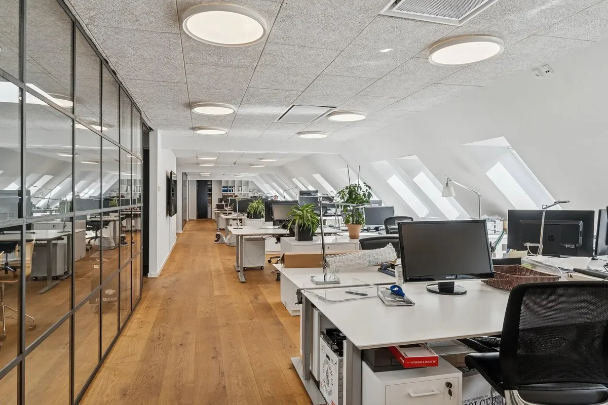 Kontor med højloft og gul trægulv, hvor flere skriveborde og computere er placeret langs væggen og midten af rummet. Rundt