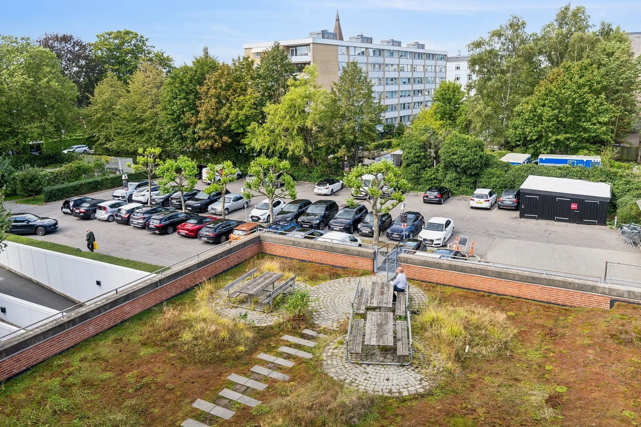 Forskudte parkeringspladser med biler og grønne haver samt et stort højhus i baggrunden