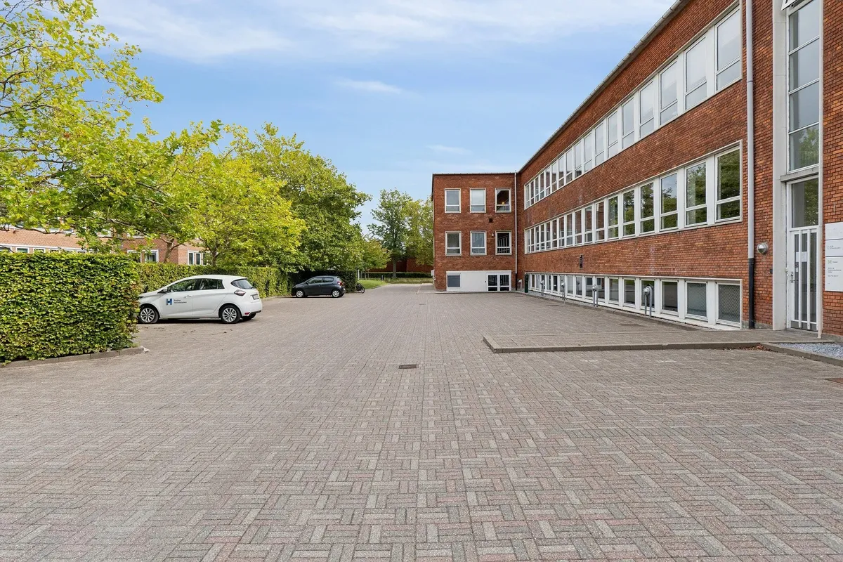 Store parkeringsplads med to biler parkeret og en række vinduer i en bygning i rød mursten og hvide vindueskarme