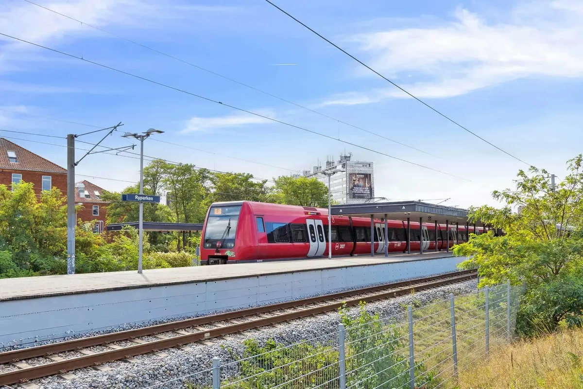 En rødt tog står stille på en station med en lang platform og grønne buske og træer i baggrunden