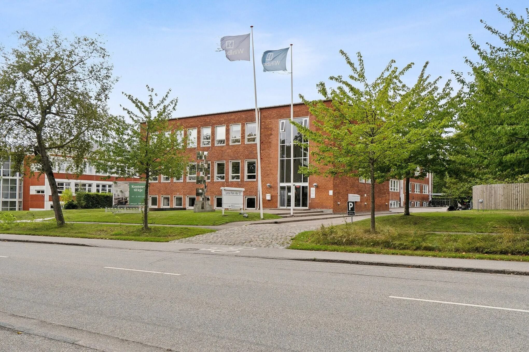 Bygning med to flag, der står i en park med grønne træer og buske på hver side af en bred asfaltvej