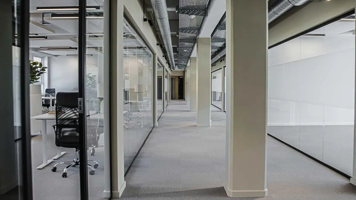 Moderne kontor med glasvægge, gulvtæppe og kontormøbler, hvoraf en hvid skrivebord med grøn plante