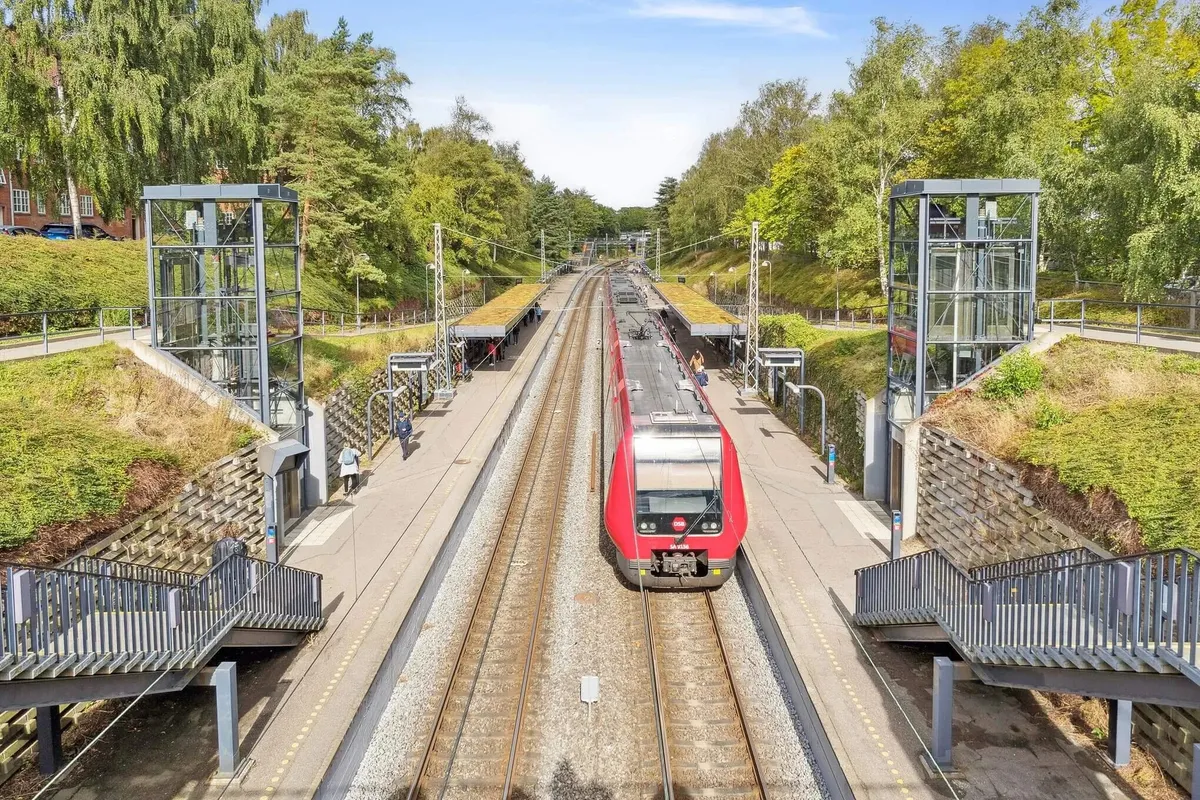 To personer går på perronen ved en rødt tog og flere mennesker står på perronens anden side uden for en glaskurv og to