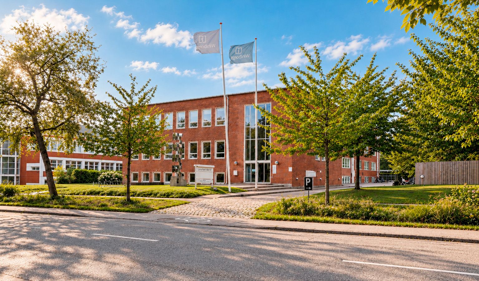 Kontorbygning med store vinduer, flag og parkeringsskilt. Solskin og grønne træer. Stenbro og skulptur. Parkområde med græsplæne. Bygningen er gul og rødt tegl.
