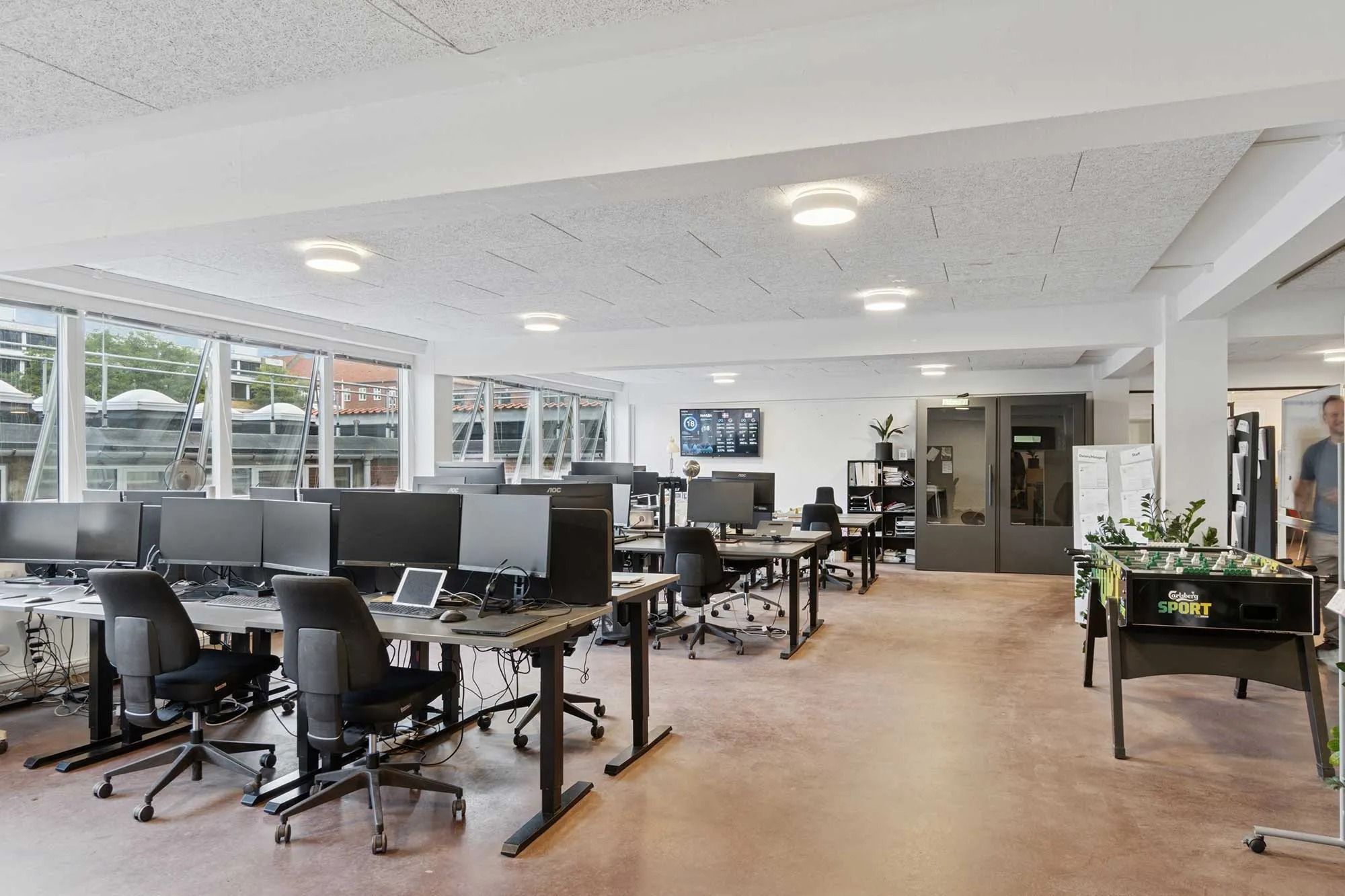 Kontor med flere skriveborde, hver med computer, stol og små plante. En stor vinduesfront giver indsigt i byen. Et