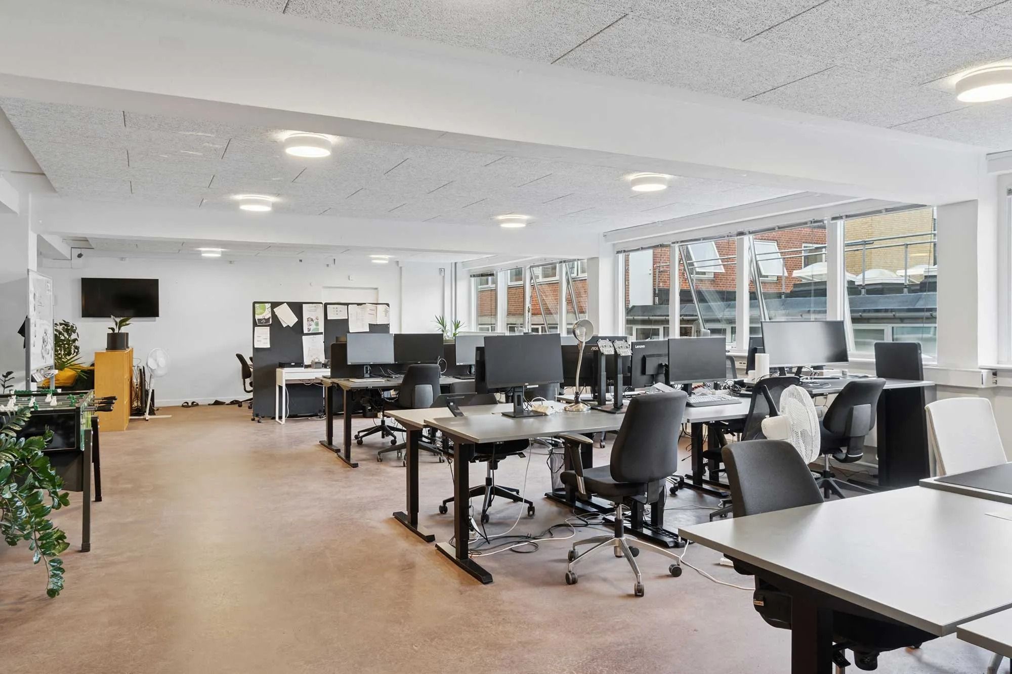 Moderne kontor med høje vinduer, betongulve og lyse lofter. Bord og stol er ordnet i rækker med computere, og gulvet er