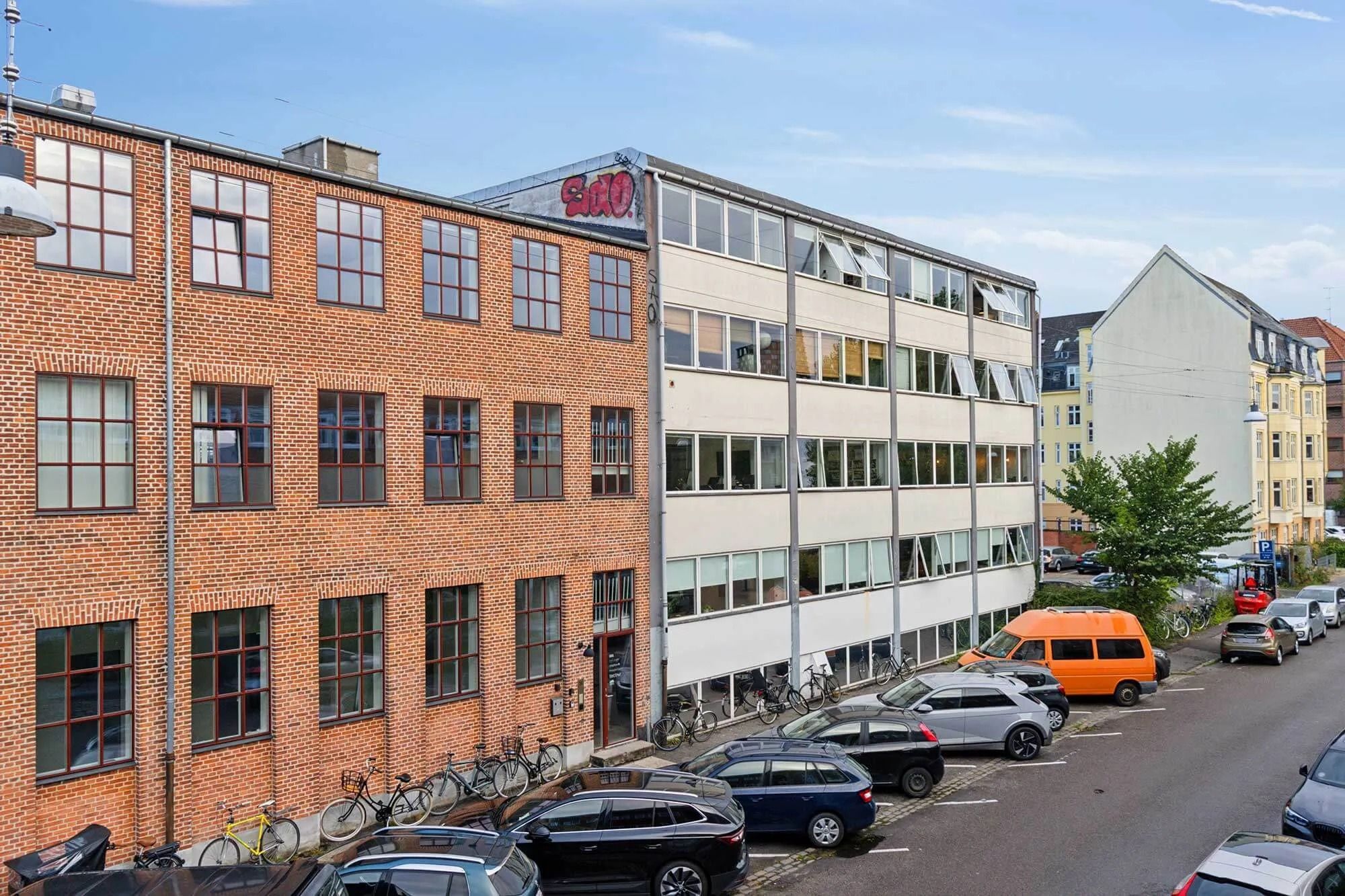 Kontorbyggeri med to rækker røde mursten og hvide vindueskarme, flere biler og en orange varevogn parkeret